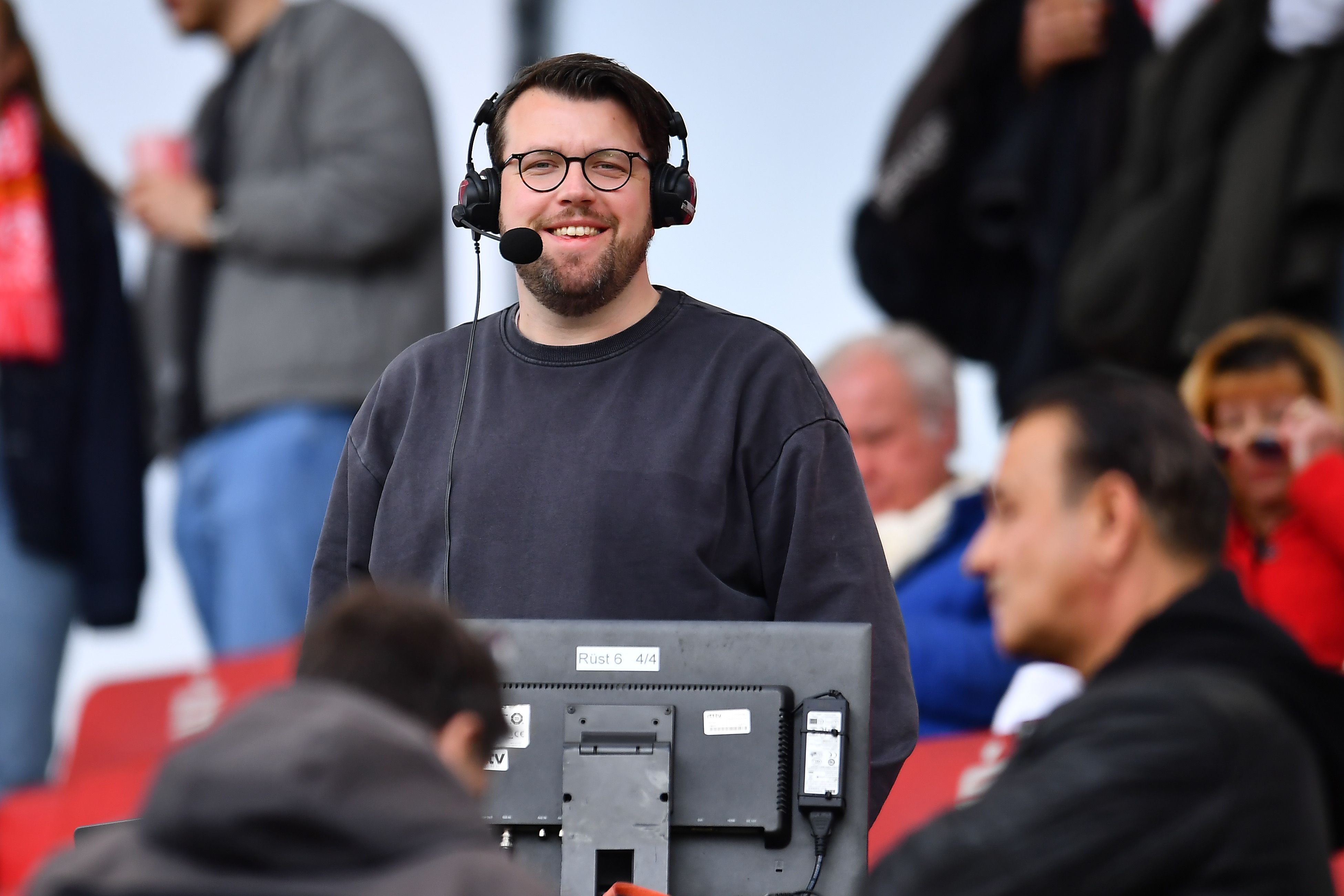 Christian Straßburger steht mit Headset am Kommentatorenplatz und lacht in die Kamera.