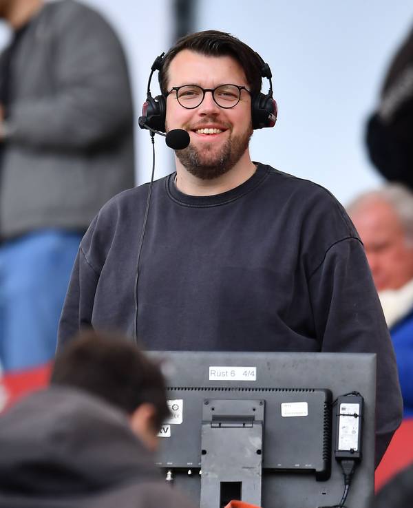 Christian Straßburger steht mit Headset am Kommentatorenplatz und lacht in die Kamera.