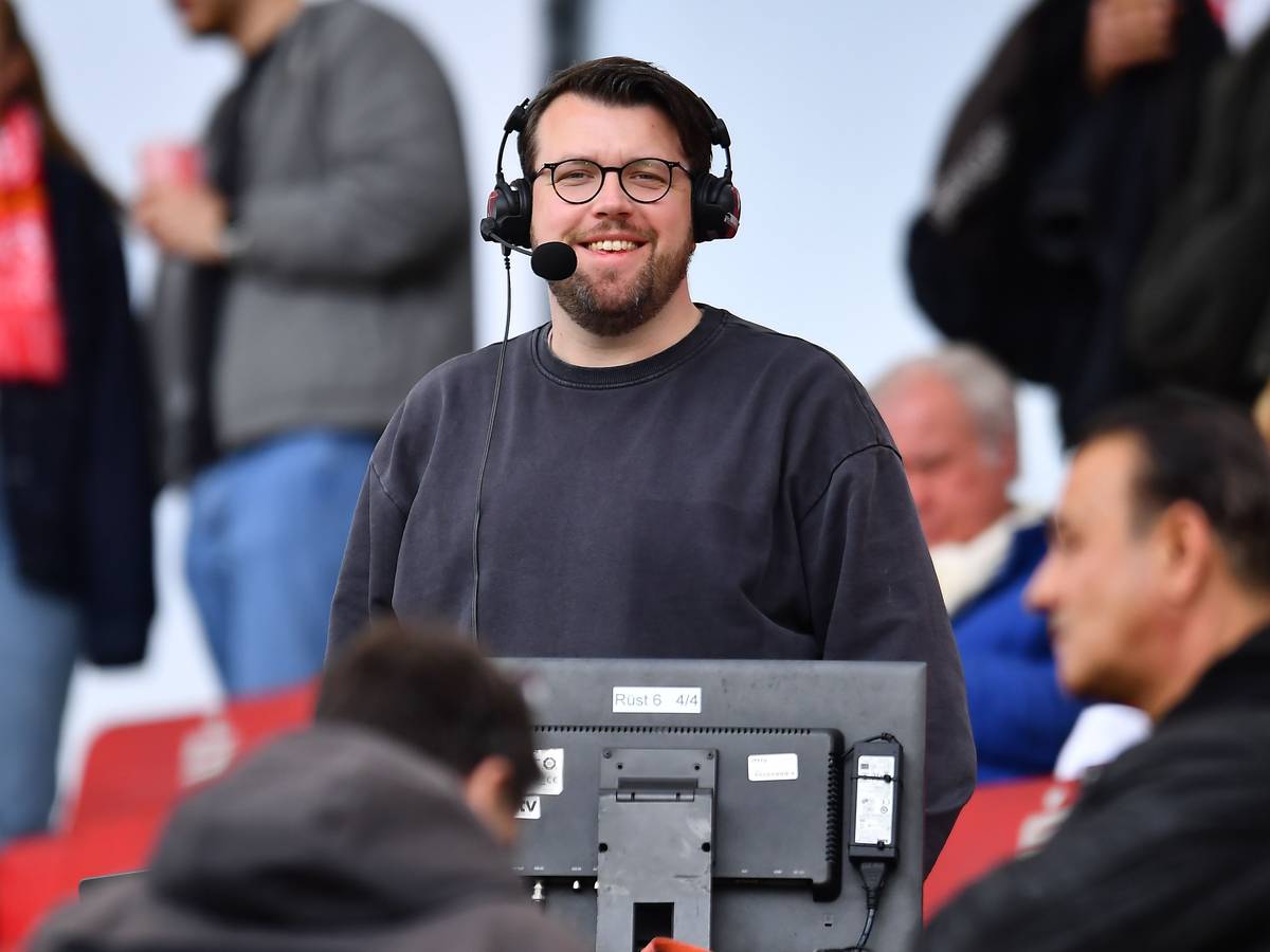 Christian Straßburger steht mit Headset am Kommentatorenplatz und lacht in die Kamera.