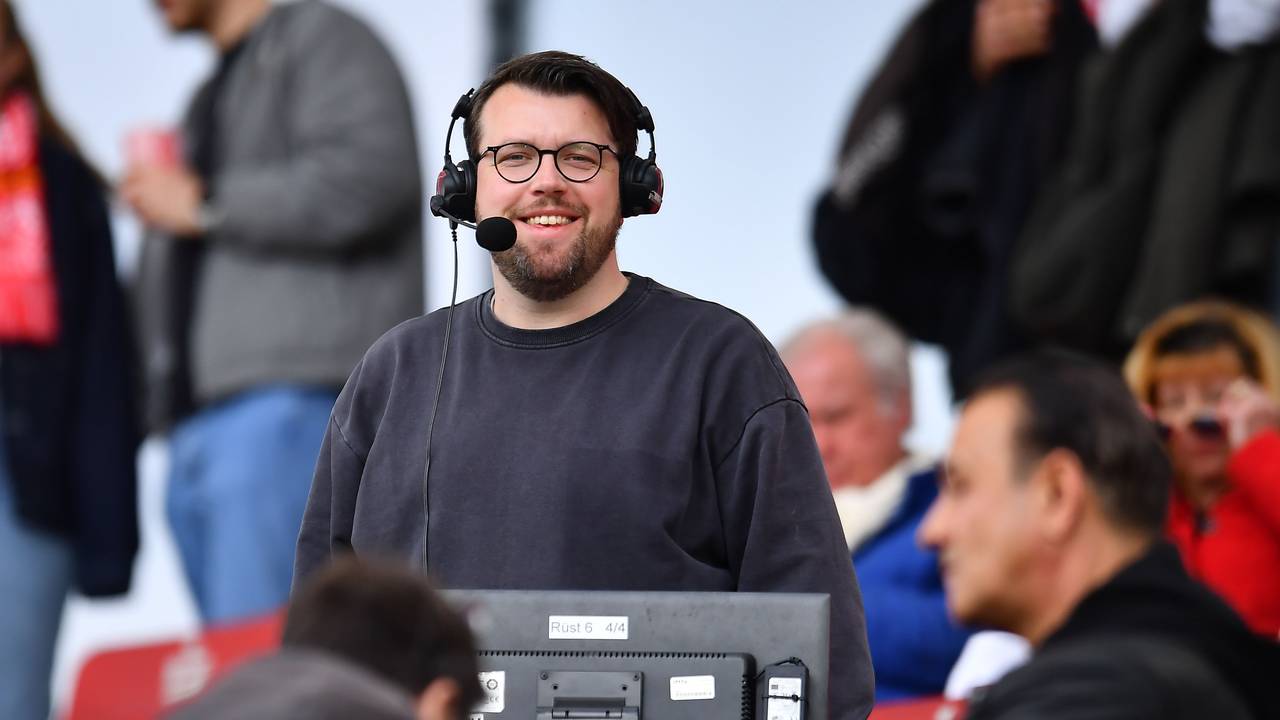 Christian Straßburger steht mit Headset am Kommentatorenplatz und lacht in die Kamera.