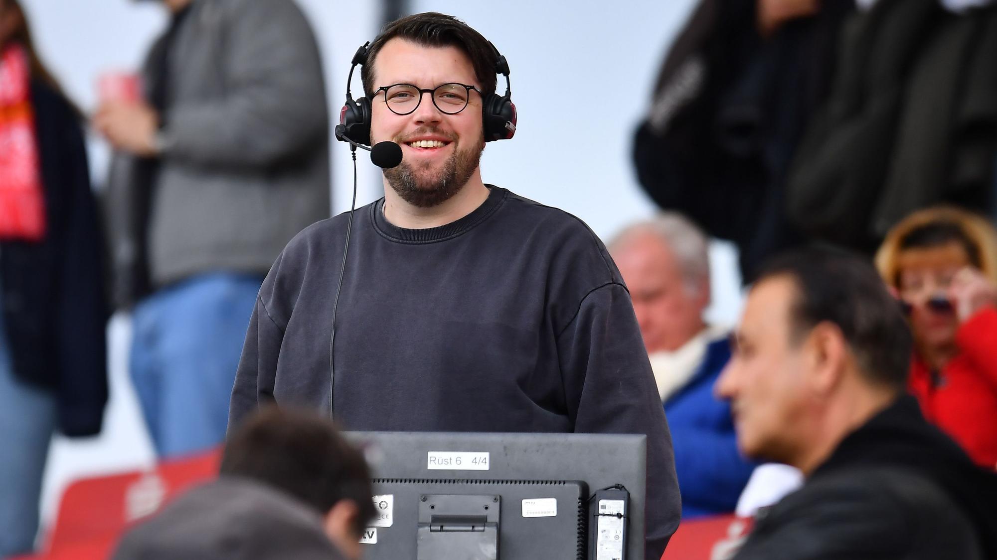 Christian Straßburger steht mit Headset am Kommentatorenplatz und lacht in die Kamera.