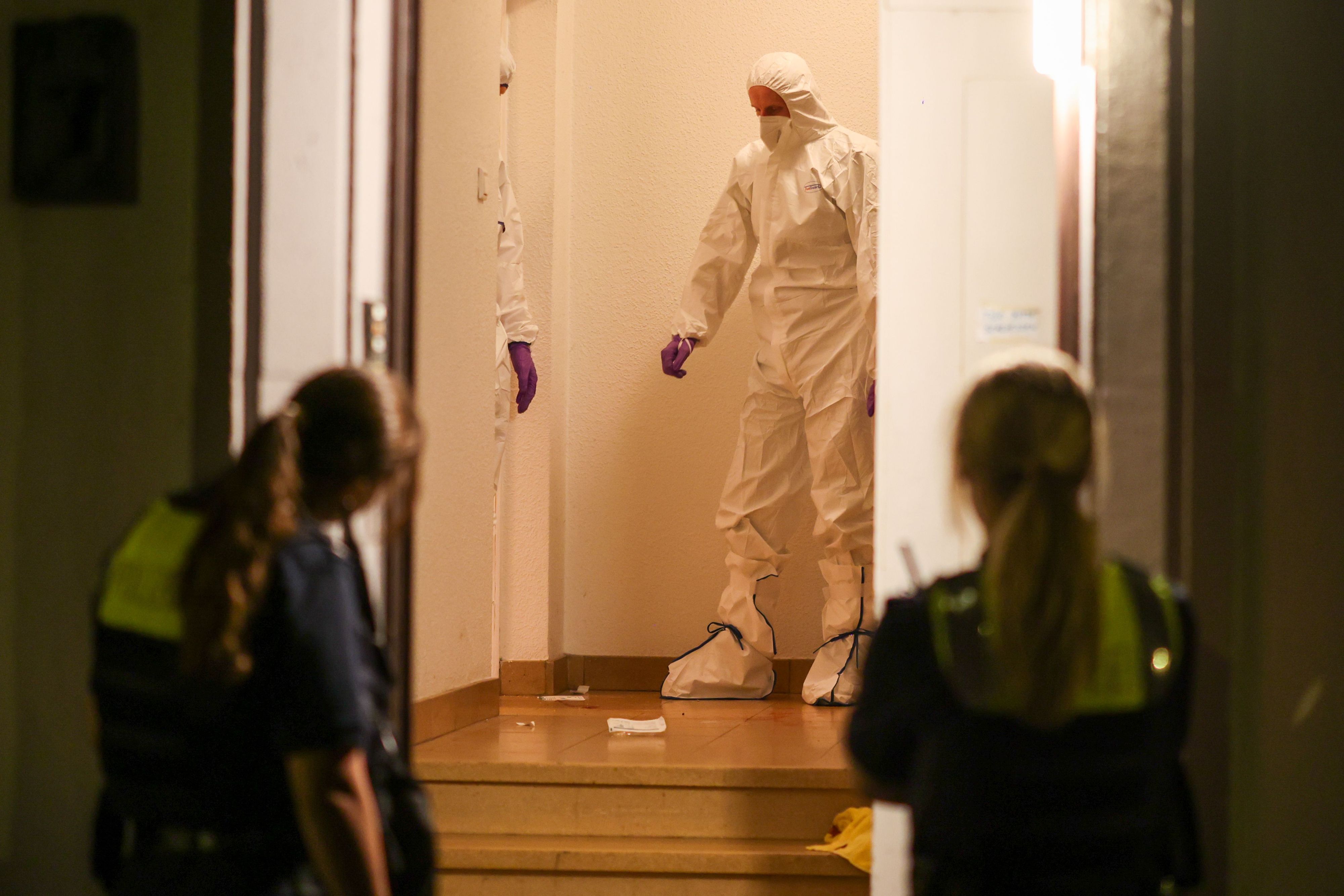 Zwei Beamte der Spurensicherung in Spurensicherungsanzügen stehen im Treppenhaus eines Mehrfamilienhauses und gucken auf etwas, das am Boden liegt. Vor der offenen Haustür werden sie von zwei Polizistinnen beobachtet.