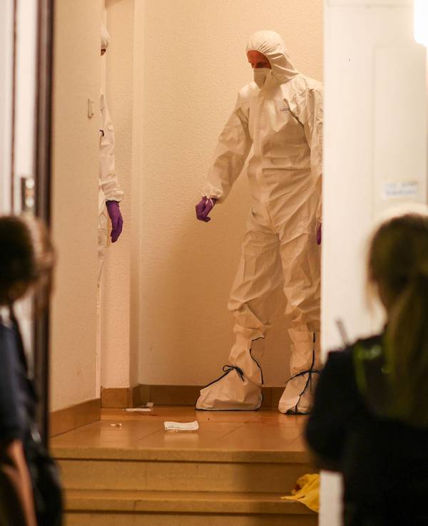 Zwei Beamte der Spurensicherung in Spurensicherungsanzügen stehen im Treppenhaus eines Mehrfamilienhauses und gucken auf etwas, das am Boden liegt. Vor der offenen Haustür werden sie von zwei Polizistinnen beobachtet.