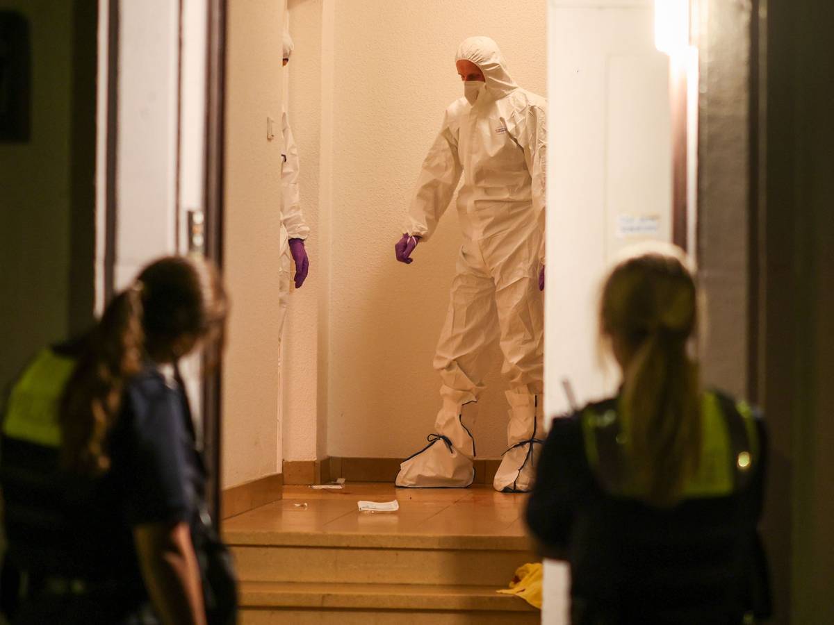 Zwei Beamte der Spurensicherung in Spurensicherungsanzügen stehen im Treppenhaus eines Mehrfamilienhauses und gucken auf etwas, das am Boden liegt. Vor der offenen Haustür werden sie von zwei Polizistinnen beobachtet.