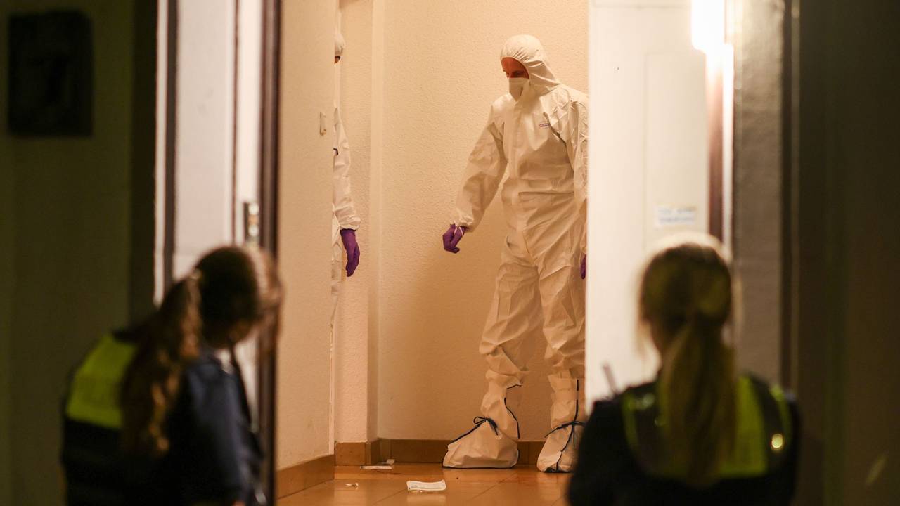Zwei Beamte der Spurensicherung in Spurensicherungsanzügen stehen im Treppenhaus eines Mehrfamilienhauses und gucken auf etwas, das am Boden liegt. Vor der offenen Haustür werden sie von zwei Polizistinnen beobachtet.