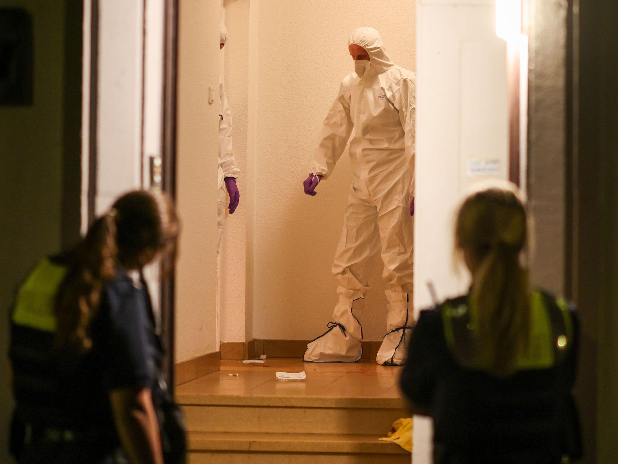 Zwei Beamte der Spurensicherung in Spurensicherungsanzügen stehen im Treppenhaus eines Mehrfamilienhauses und gucken auf etwas, das am Boden liegt. Vor der offenen Haustür werden sie von zwei Polizistinnen beobachtet.