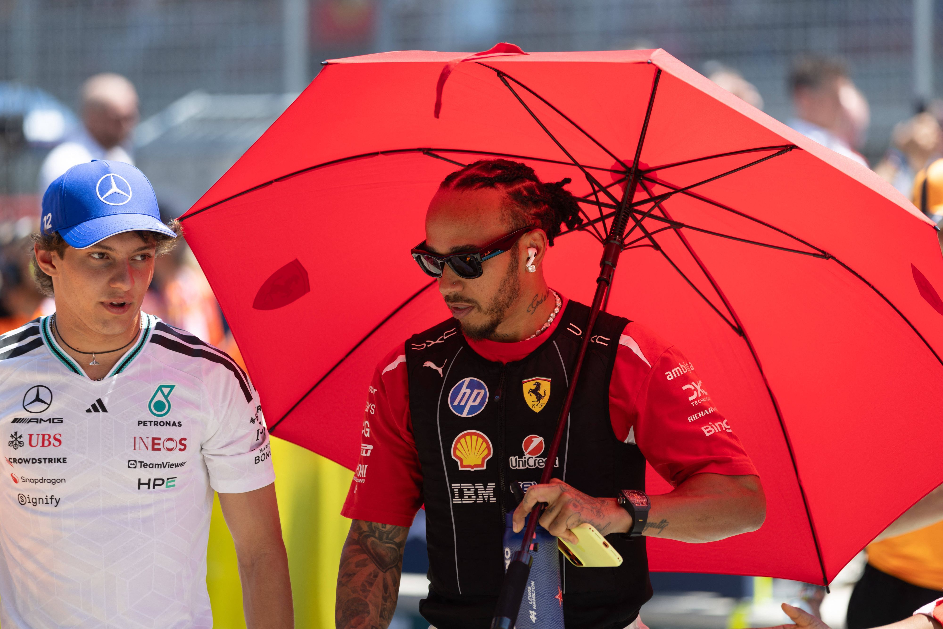 Lewis Hamilton schützt sich unter einem roten Schirm vo der Sonne, neben ihm geht Kimi Antonelli.