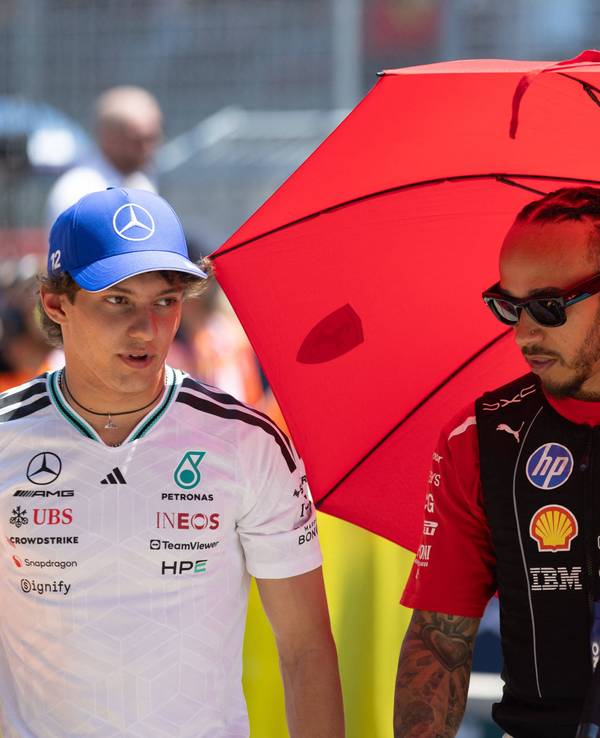 Lewis Hamilton schützt sich unter einem roten Schirm vo der Sonne, neben ihm geht Kimi Antonelli.