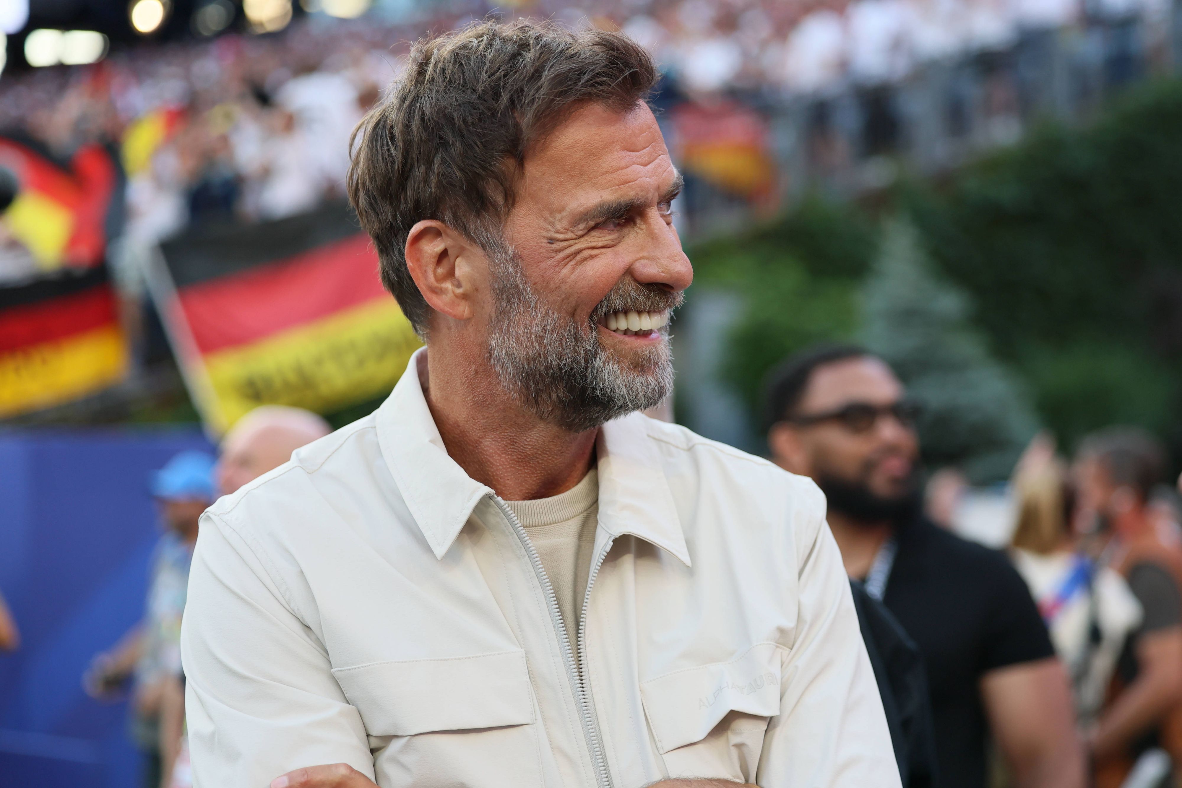 Jürgen Klopp steht im Stadion und lächelt. Hinter ihm ist verschwommen eine Deutschland-Flagge von Fans zu sehen.