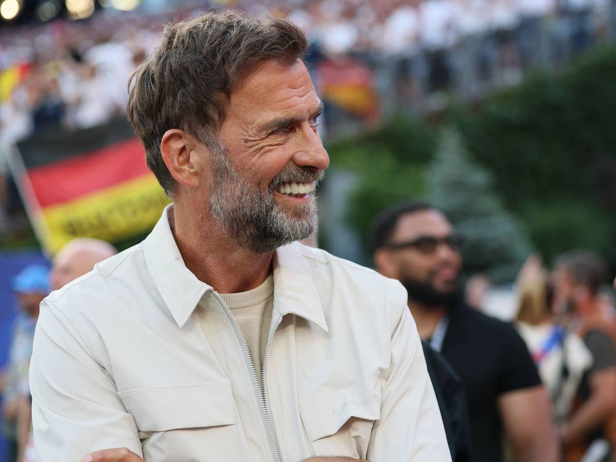 Jürgen Klopp steht im Stadion und lächelt. Hinter ihm ist verschwommen eine Deutschland-Flagge von Fans zu sehen.