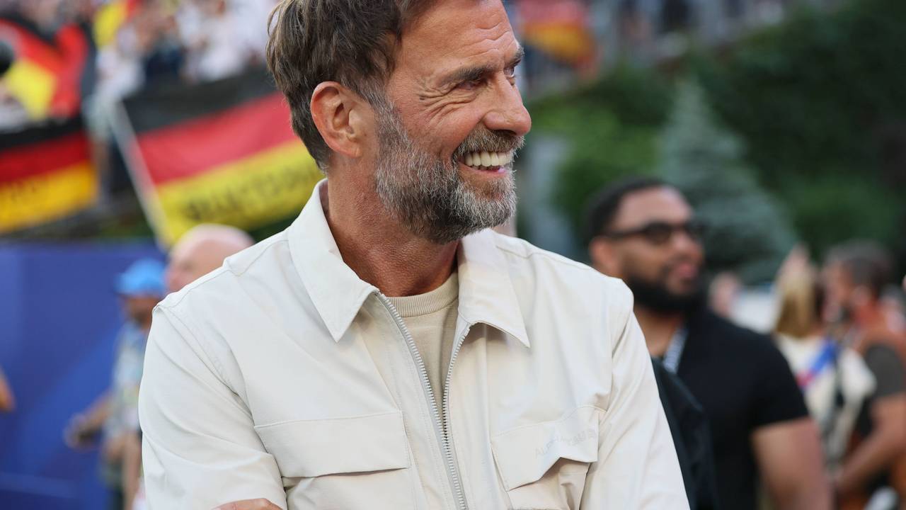 Jürgen Klopp steht im Stadion und lächelt. Hinter ihm ist verschwommen eine Deutschland-Flagge von Fans zu sehen.