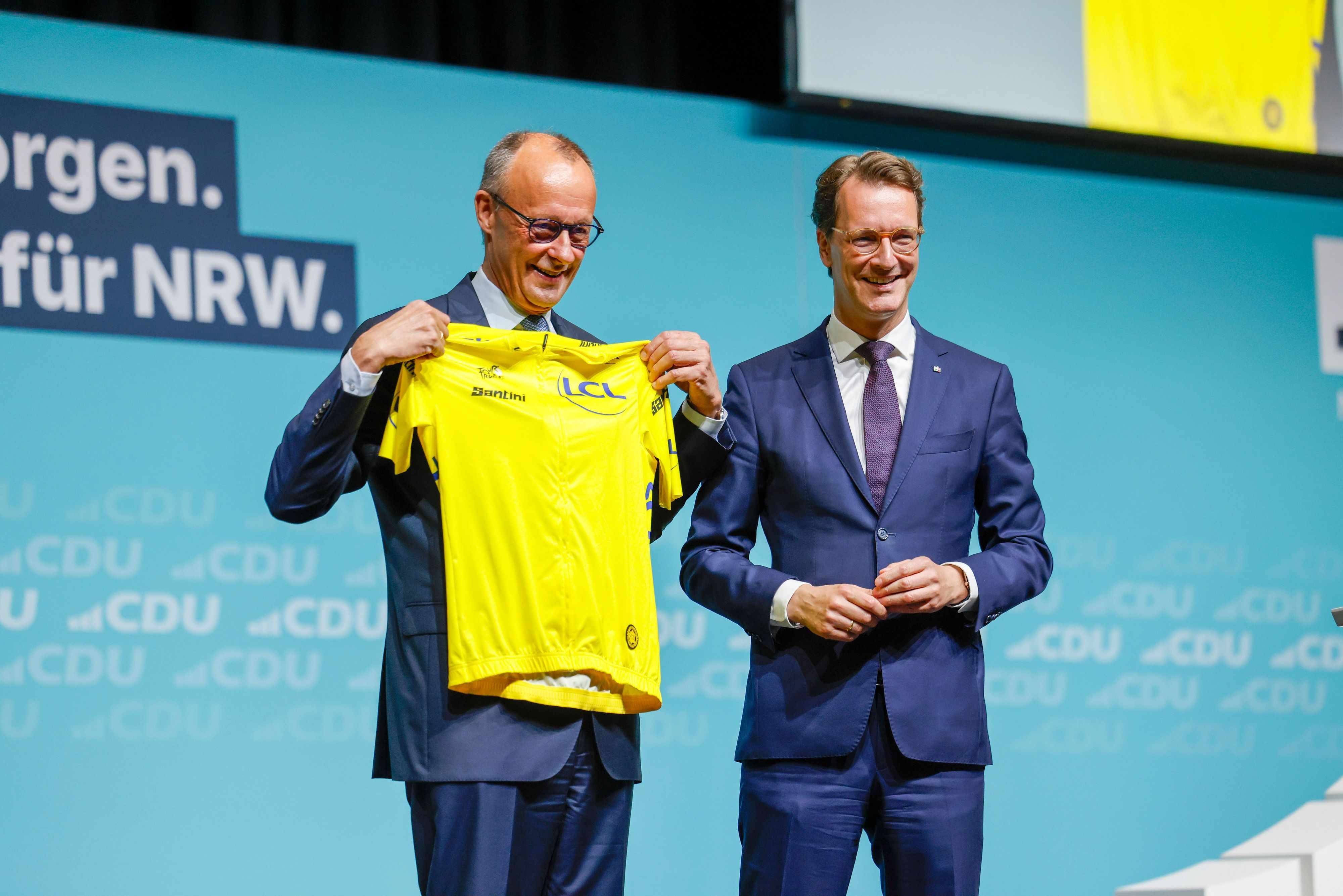 Hendrik Wüst (r., CDU), Ministerpräsident von Nordrhein-Westfalen, überreichte Bundeskanzler Friedrich Merz (CDU) beim Landesparteitag der CDU Nordrhein-Westfalen ein gelbes Rennradtrikot.
