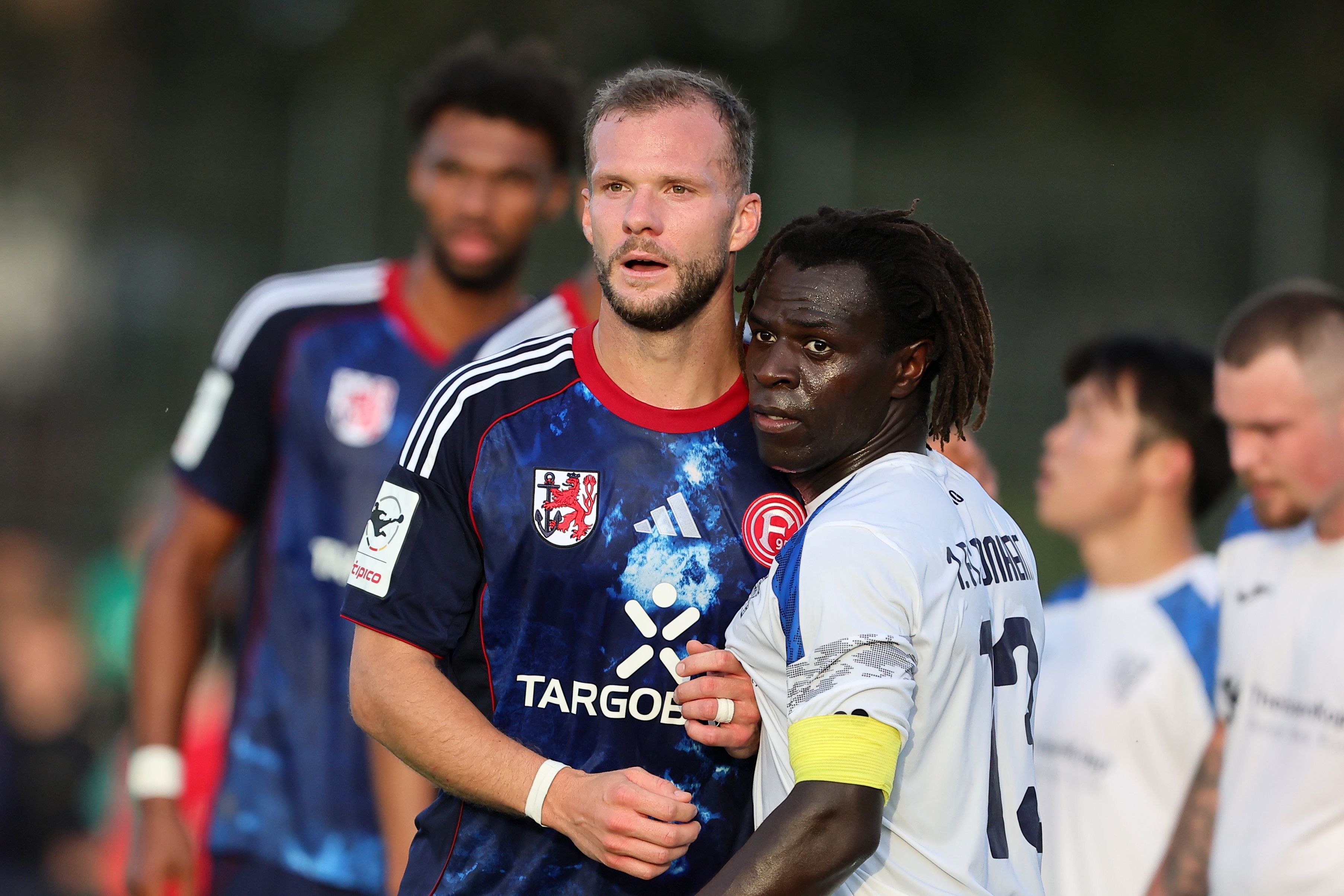 Eric Hottmann steht in einem blauen Trikot von Fortuna Düsseldorf neben seinem Gegenspieler aus Monheim.