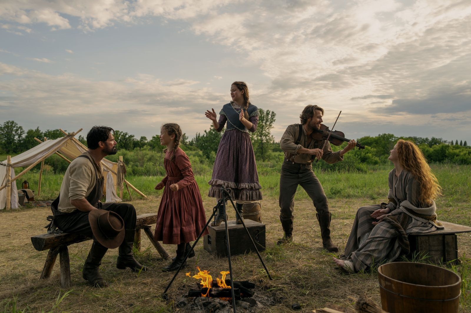 Fröhliche Zeiten in der Prärie: Schon in den Büchern griff Vater Charles Ingalls (Luke Bracey, zweiter von rechts), gerne zur Fiddle und erfreute damit seine Töchter Laura (Alice Halsey, zweite von links), Mary (Skywalker Hughes, Mitte), Ehefrau Caroline (Crosby Fitzgerald) und Familienfreund Mr. Edwards (Warren Christie). (Bild: Eric Zachanowich/Netflix © 2026)