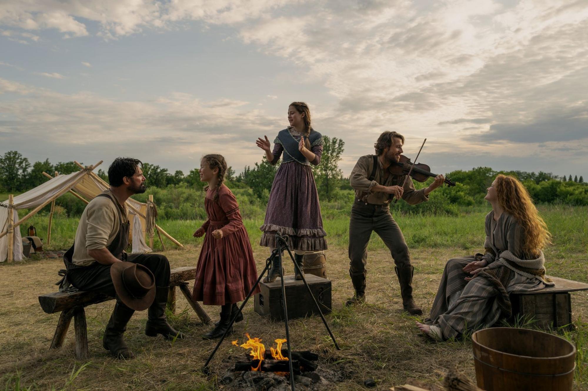 Fröhliche Zeiten in der Prärie: Schon in den Büchern griff Vater Charles Ingalls (Luke Bracey, zweiter von rechts), gerne zur Fiddle und erfreute damit seine Töchter Laura (Alice Halsey, zweite von links), Mary (Skywalker Hughes, Mitte), Ehefrau Caroline (Crosby Fitzgerald) und Familienfreund Mr. Edwards (Warren Christie). (Bild: Eric Zachanowich/Netflix © 2026)