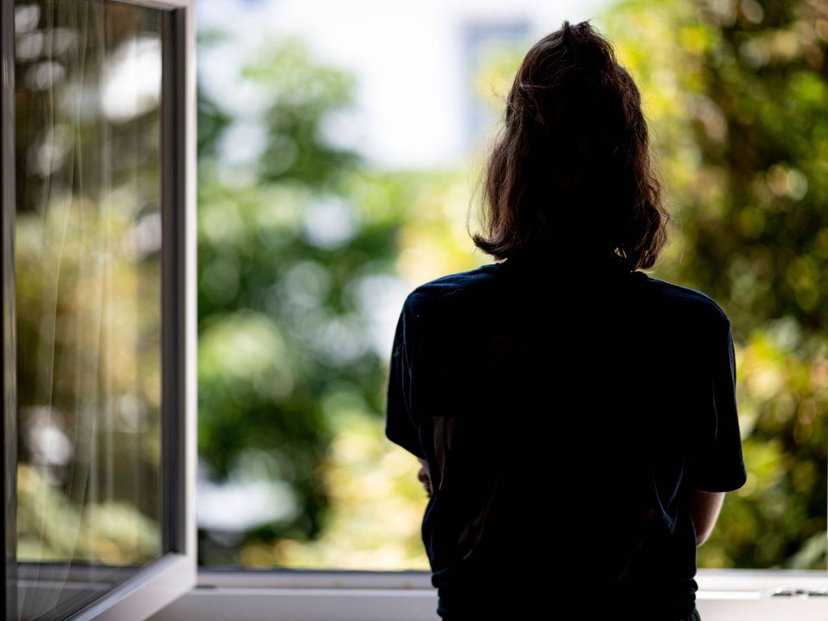 Eine junge Frau steht in ihrer Wohnung an einem Fenster.