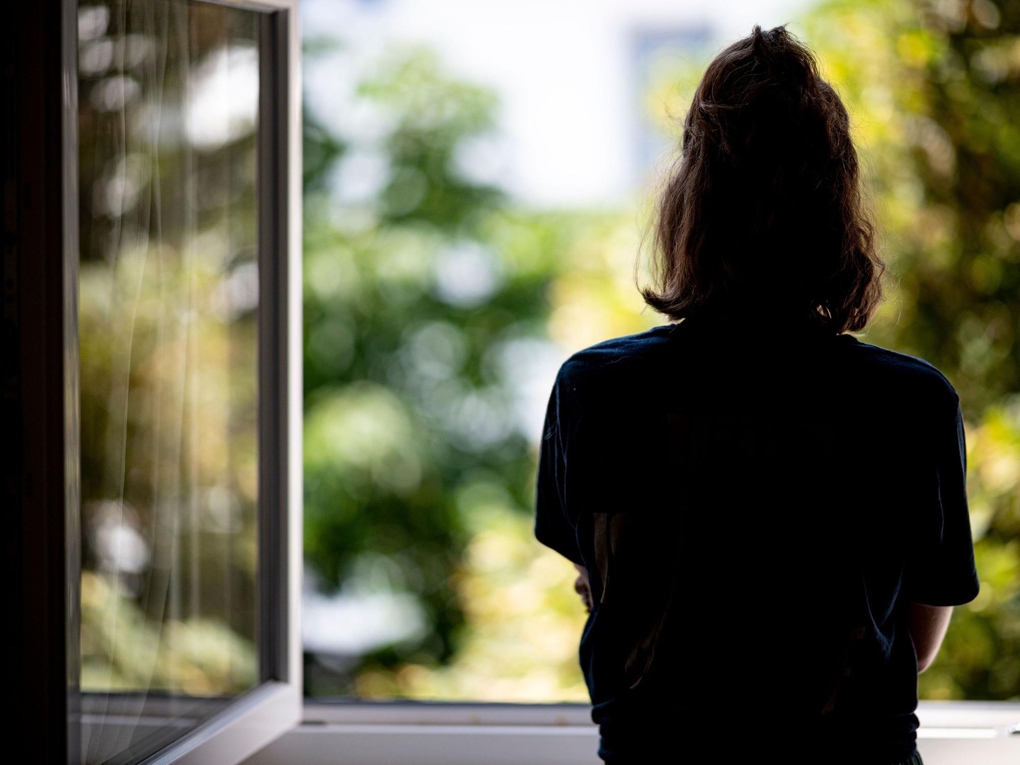 Eine junge Frau steht in ihrer Wohnung an einem Fenster.