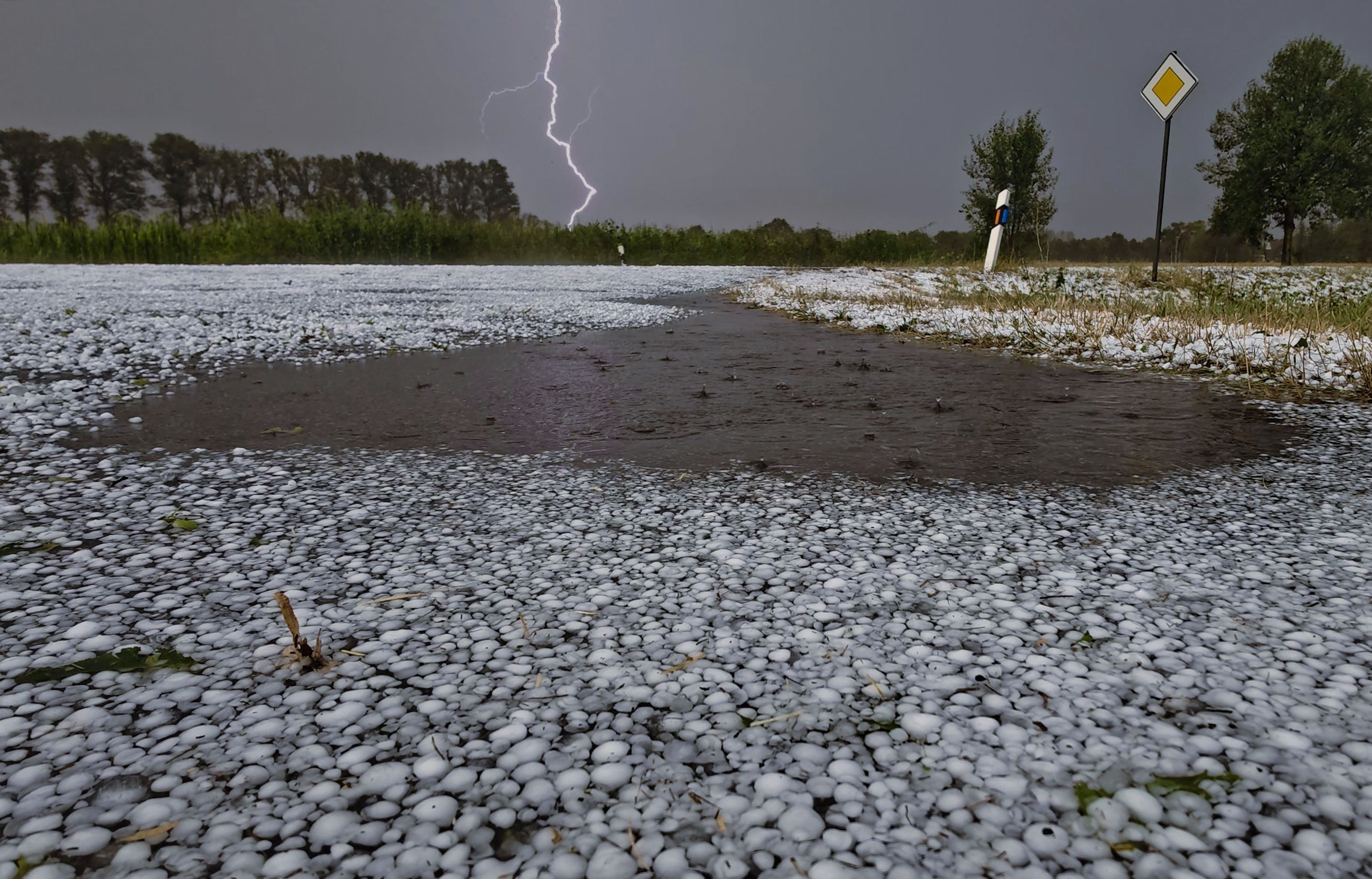 Schwere Unwetter sind über Teilen Niedersachsens niedergegangen. Hagelkörner liegen auf einer Fahrbahn bei Wagenfeld in Niedersachsen, während sich ein Blitz im Hintergrund entlädt.