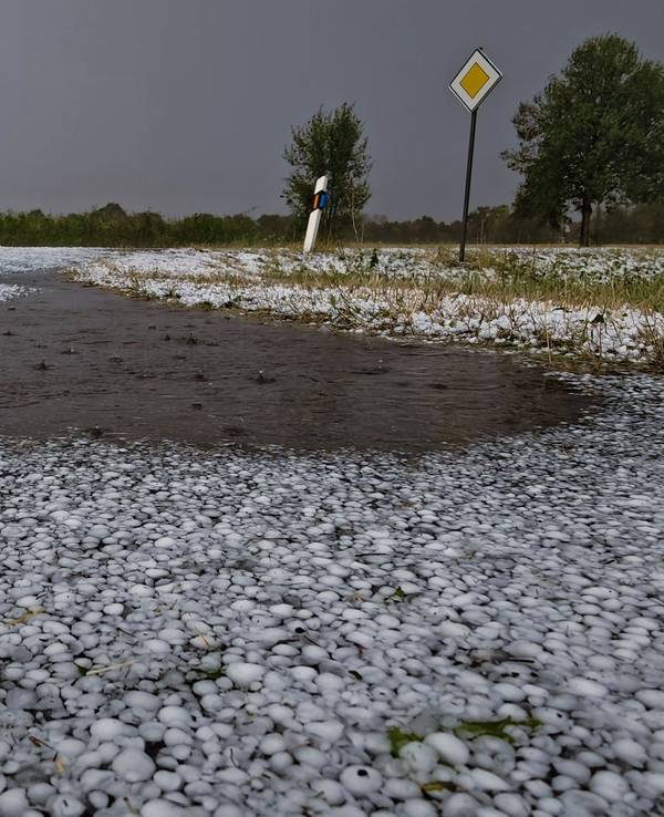 Schwere Unwetter sind über Teilen Niedersachsens niedergegangen. Hagelkörner liegen auf einer Fahrbahn bei Wagenfeld in Niedersachsen, während sich ein Blitz im Hintergrund entlädt.