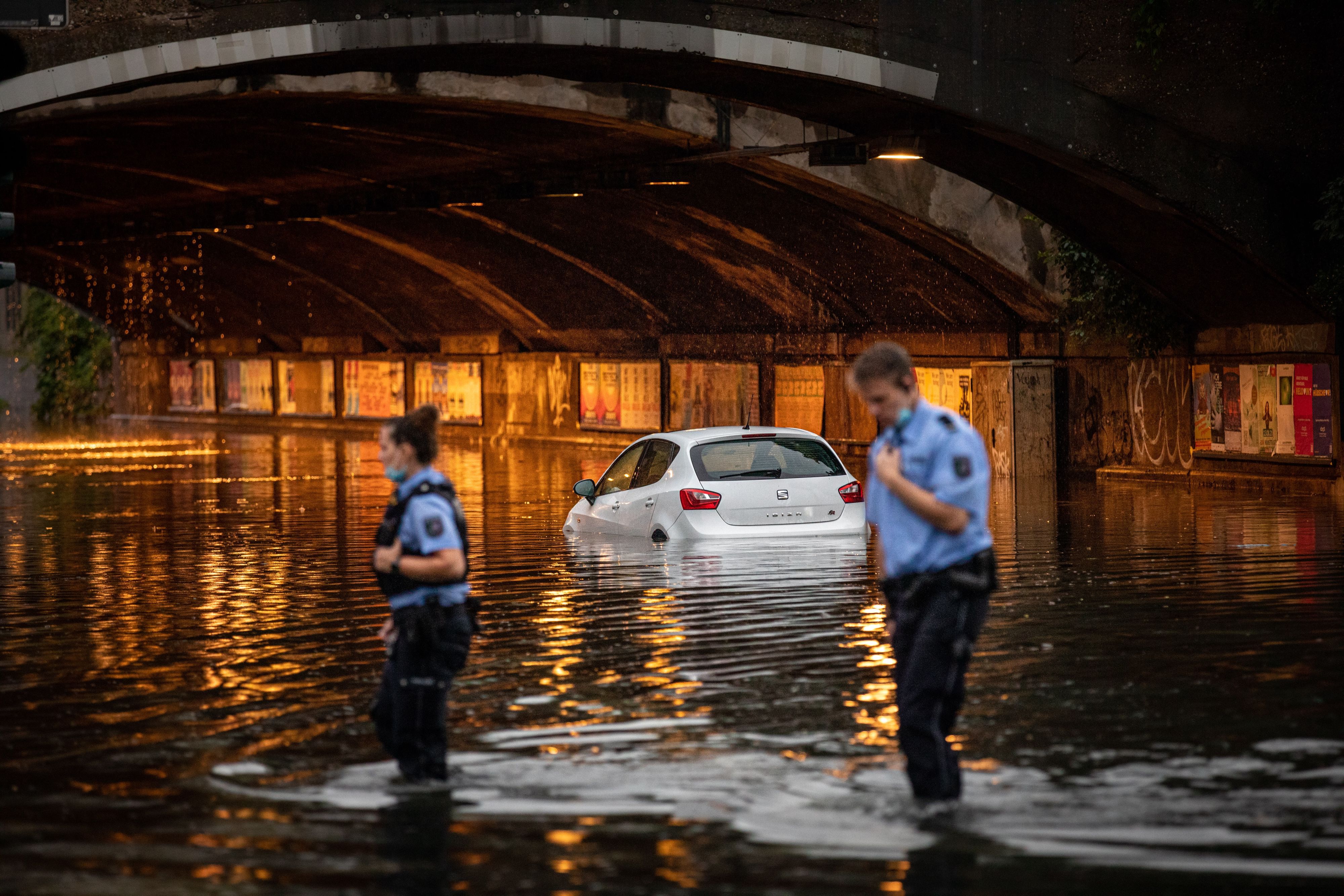 Eine Unterführung ist überflutet, ein Auto steht bis zur Hälfte im Wasser, einem Polizist und einer Polizistin reicht das Wasser fast bis zu den Knien.