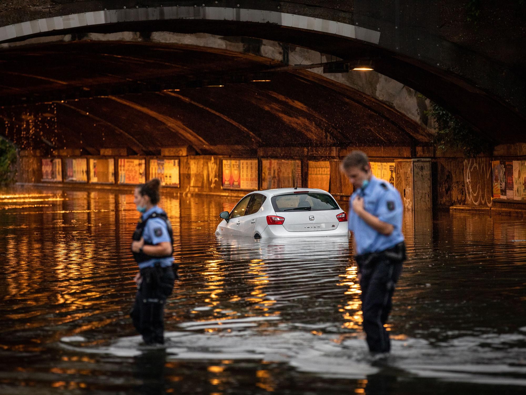 Eine Unterführung ist überflutet, ein Auto steht bis zur Hälfte im Wasser, einem Polizist und einer Polizistin reicht das Wasser fast bis zu den Knien.