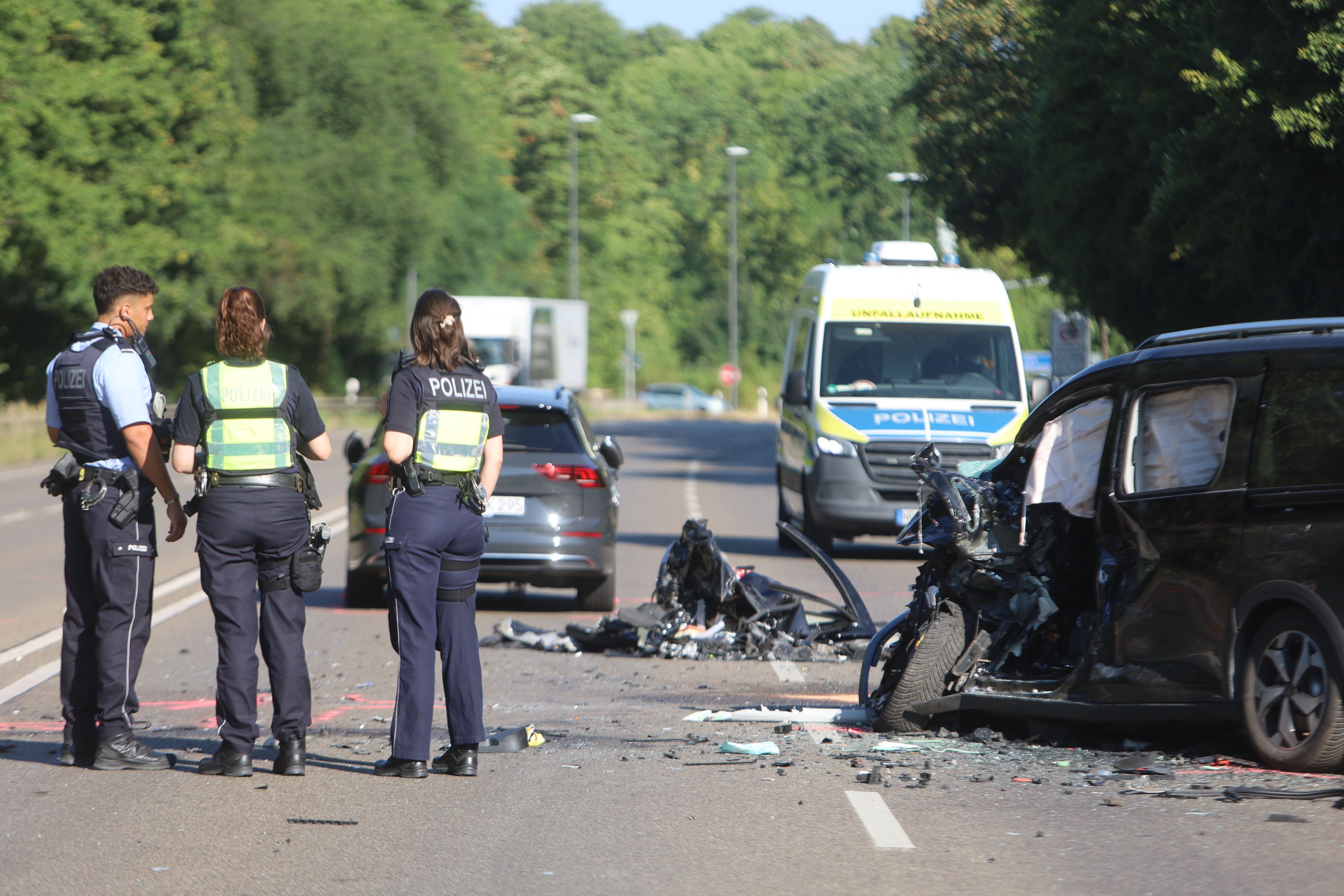 Einsatzkräfte der Polizei stehen am Mittwochmorgen (15. Juli 2026) an der Unfallstelle. Nach dem Frontalcrash liegen zahlreiche Trümmerteile auf den Fahrbahnen der Kölner Straße.