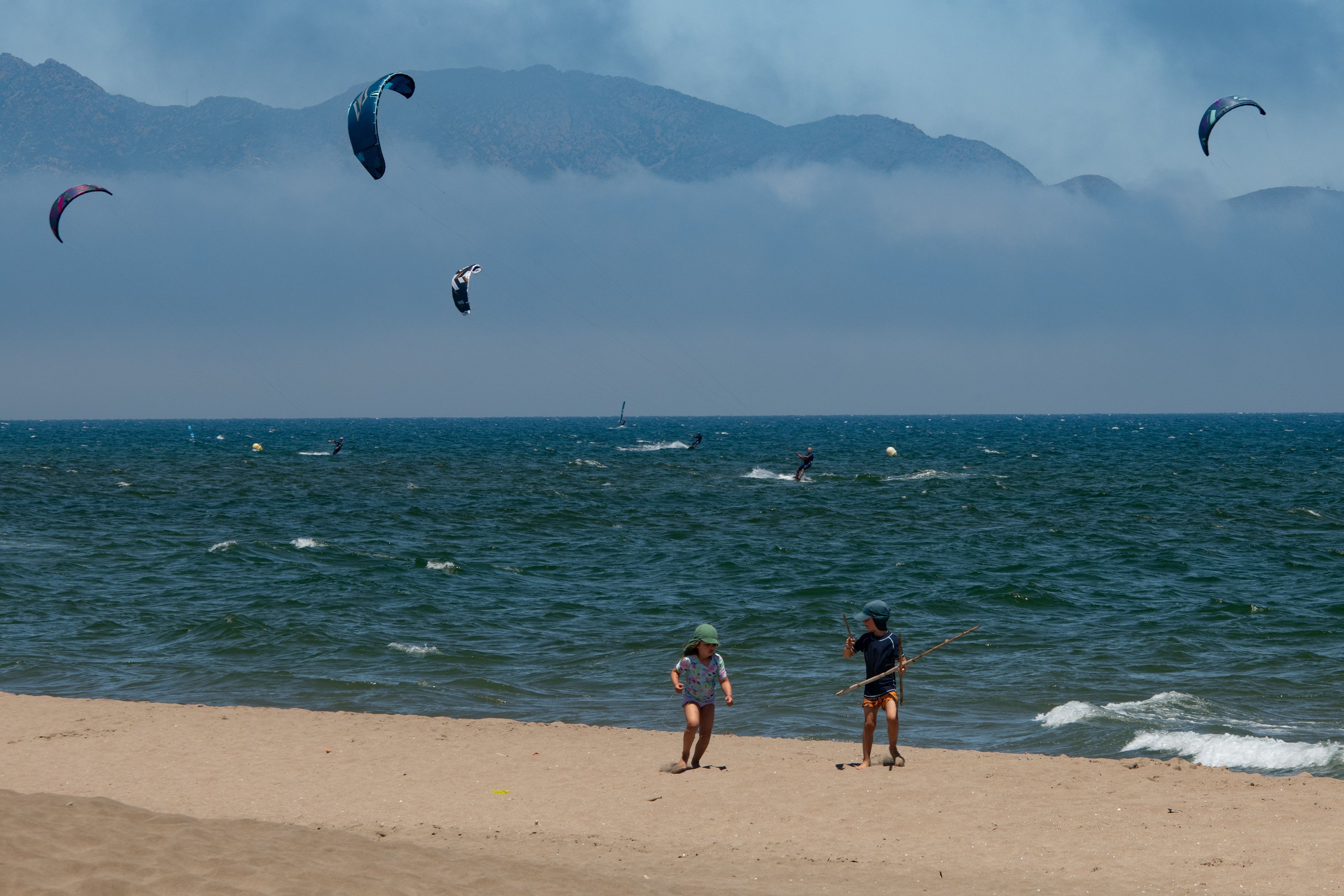 Diese Urlaubsszene ist für viele Menschen in NRW Luxus: Kinder spielen an einem warmen Sommertag am Strand, während sich Menschen im Meer beim Kitesurfen amüsieren.