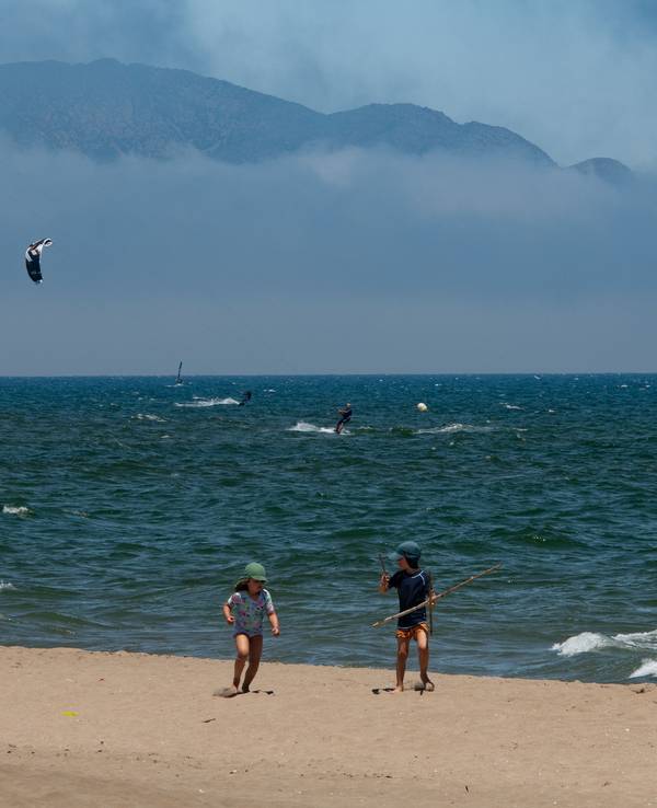 Diese Urlaubsszene ist für viele Menschen in NRW Luxus: Kinder spielen an einem warmen Sommertag am Strand, während sich Menschen im Meer beim Kitesurfen amüsieren.