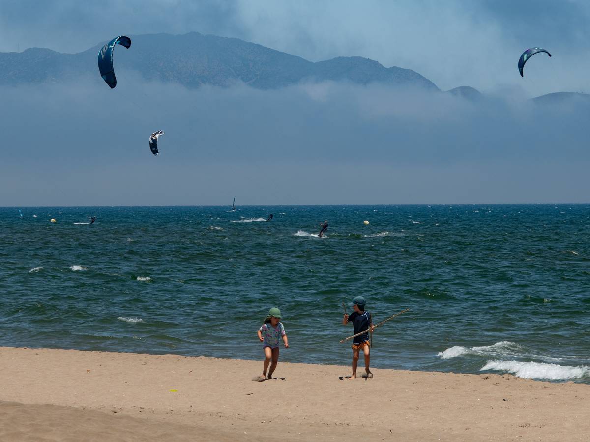 Diese Urlaubsszene ist für viele Menschen in NRW Luxus: Kinder spielen an einem warmen Sommertag am Strand, während sich Menschen im Meer beim Kitesurfen amüsieren.