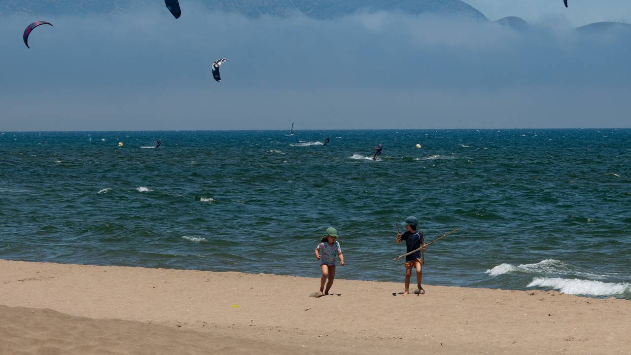 Diese Urlaubsszene ist für viele Menschen in NRW Luxus: Kinder spielen an einem warmen Sommertag am Strand, während sich Menschen im Meer beim Kitesurfen amüsieren.