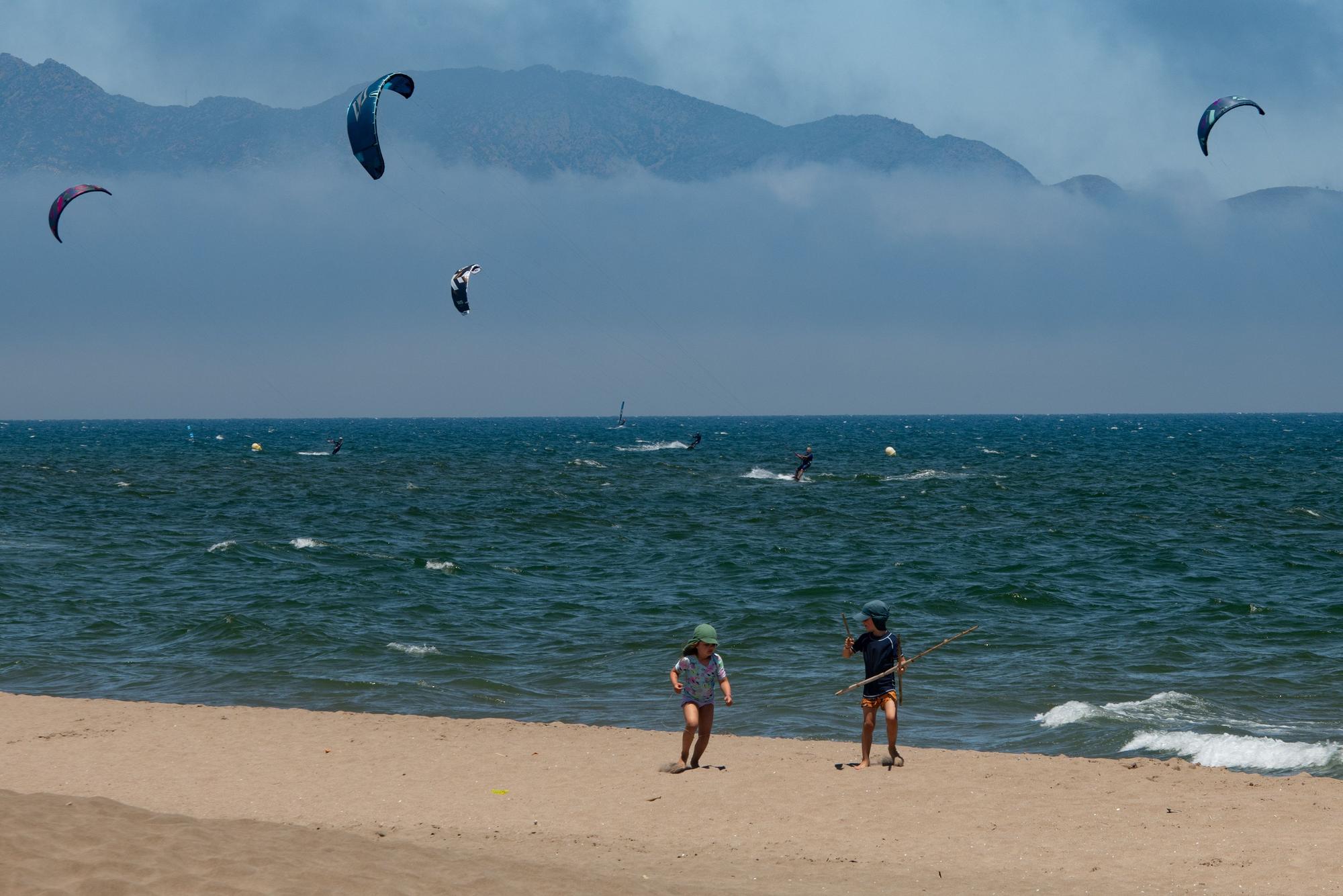Diese Urlaubsszene ist für viele Menschen in NRW Luxus: Kinder spielen an einem warmen Sommertag am Strand, während sich Menschen im Meer beim Kitesurfen amüsieren.