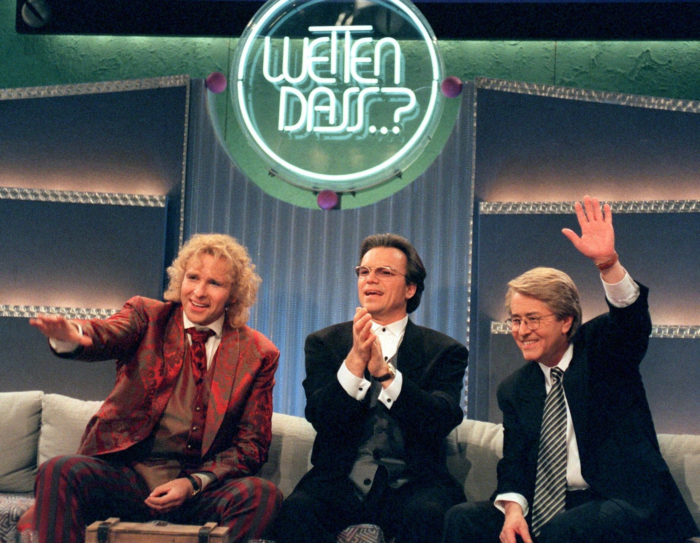 Drei schick gekleifdete Herren albern auf einem Sofa unter dem Logo von "Wetten, dass..?!" herum.