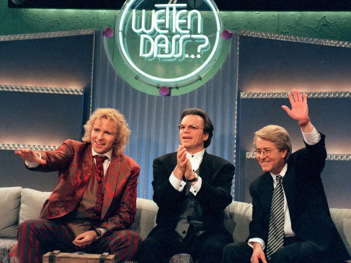 Drei schick gekleifdete Herren albern auf einem Sofa unter dem Logo von "Wetten, dass..?!" herum.