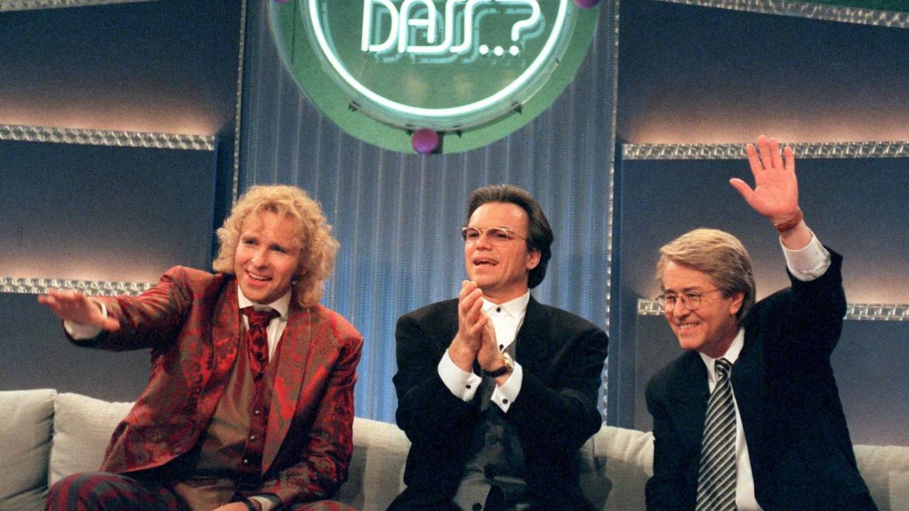 Drei schick gekleifdete Herren albern auf einem Sofa unter dem Logo von "Wetten, dass..?!" herum.