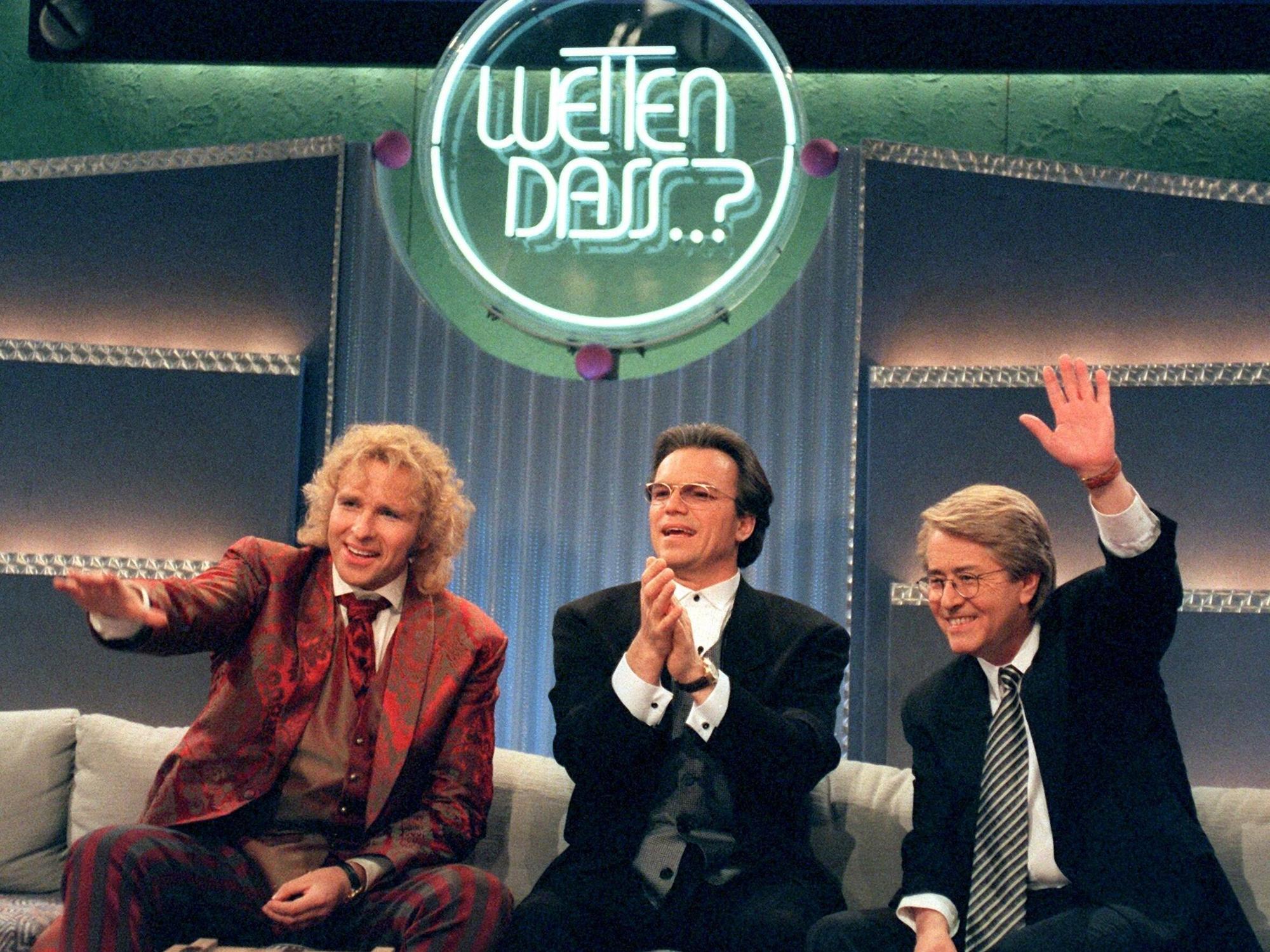 Drei schick gekleifdete Herren albern auf einem Sofa unter dem Logo von "Wetten, dass..?!" herum.