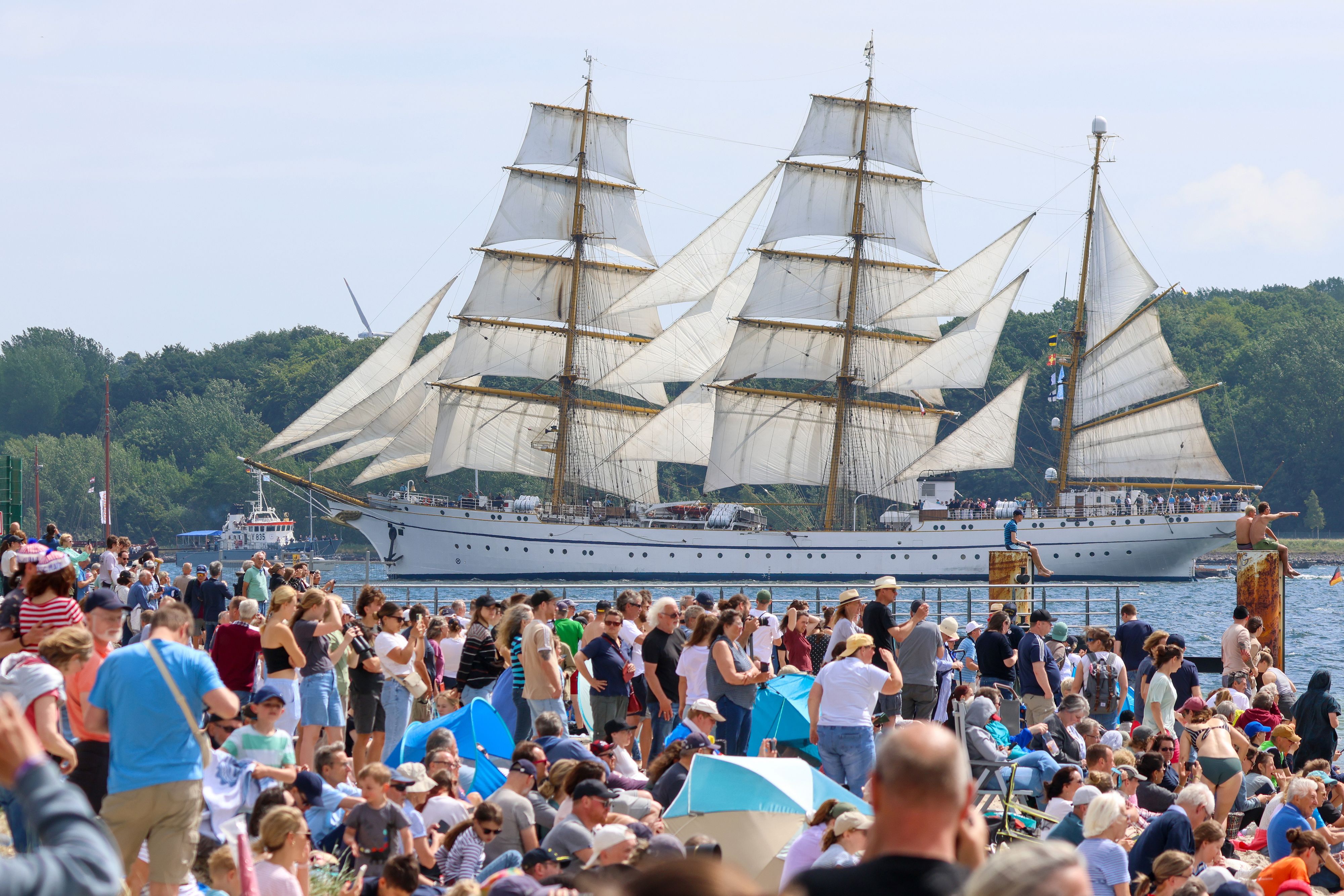 An einem Ufer drängen sich Mensch, im Hintergrund schwimmt ein dreimastiges Segelschiff.