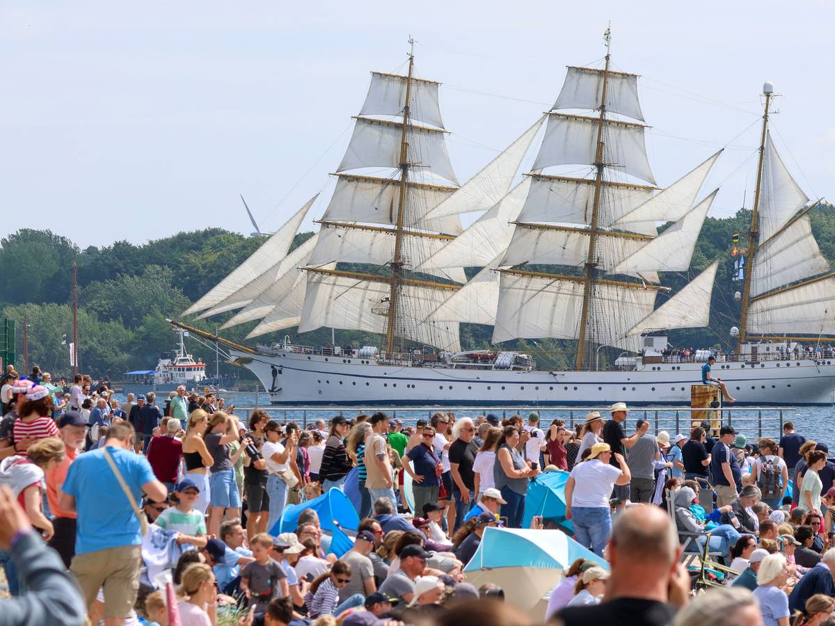 An einem Ufer drängen sich Mensch, im Hintergrund schwimmt ein dreimastiges Segelschiff.