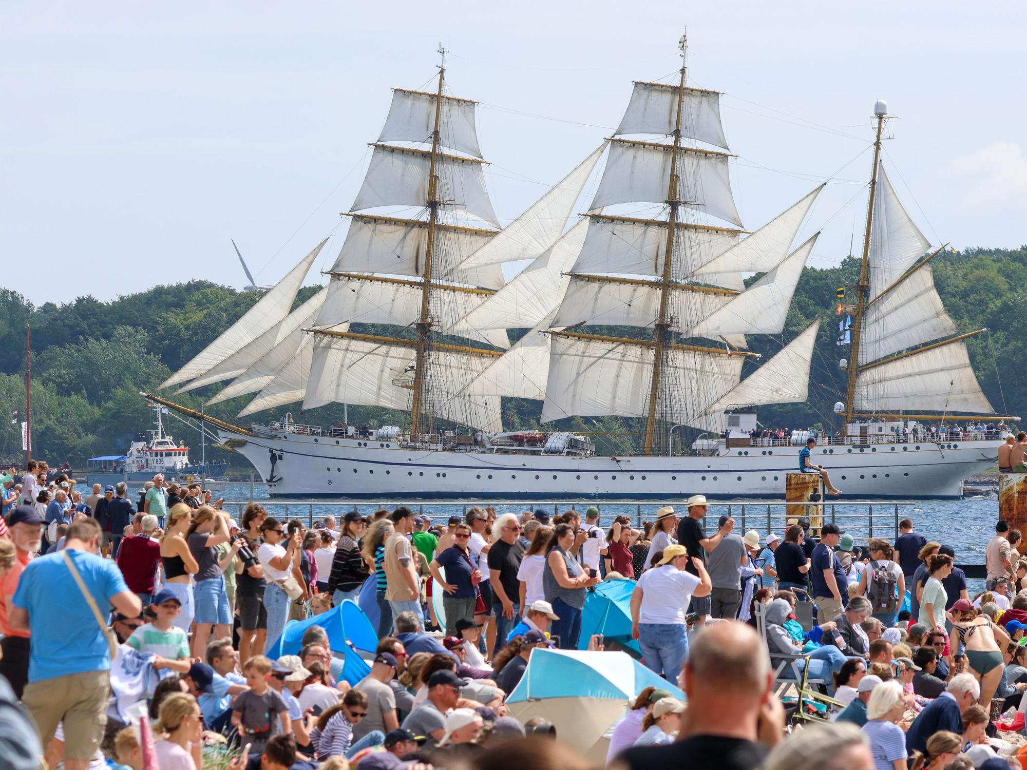 An einem Ufer drängen sich Mensch, im Hintergrund schwimmt ein dreimastiges Segelschiff.