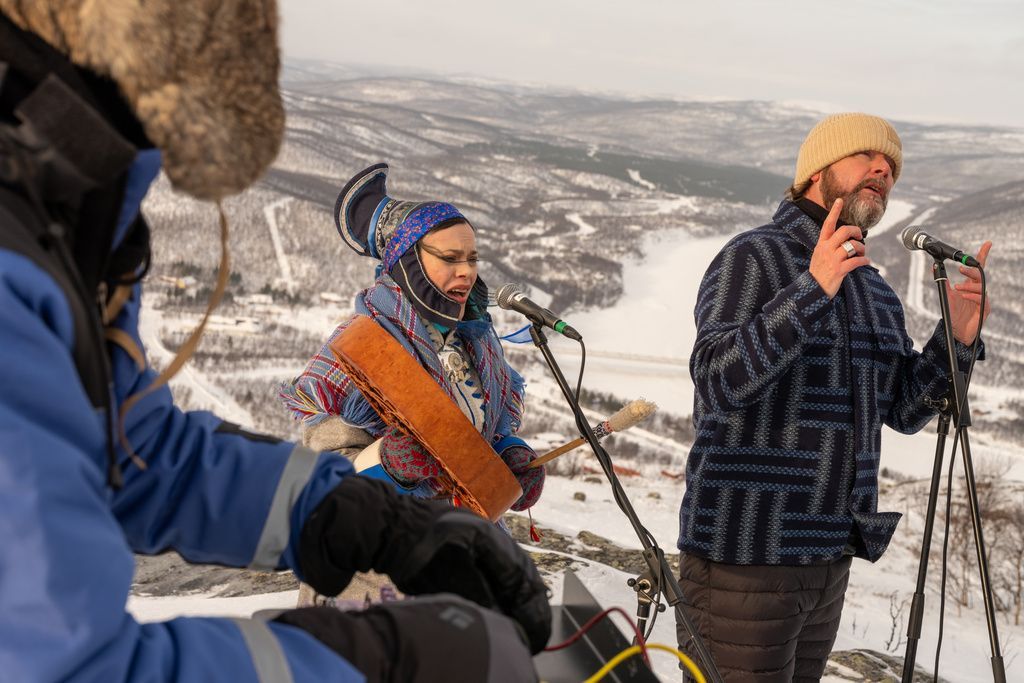 Eine Frau in nordischer Tracht und ein Man mit Mütze singen vor einer verschneiten Bergkulisse