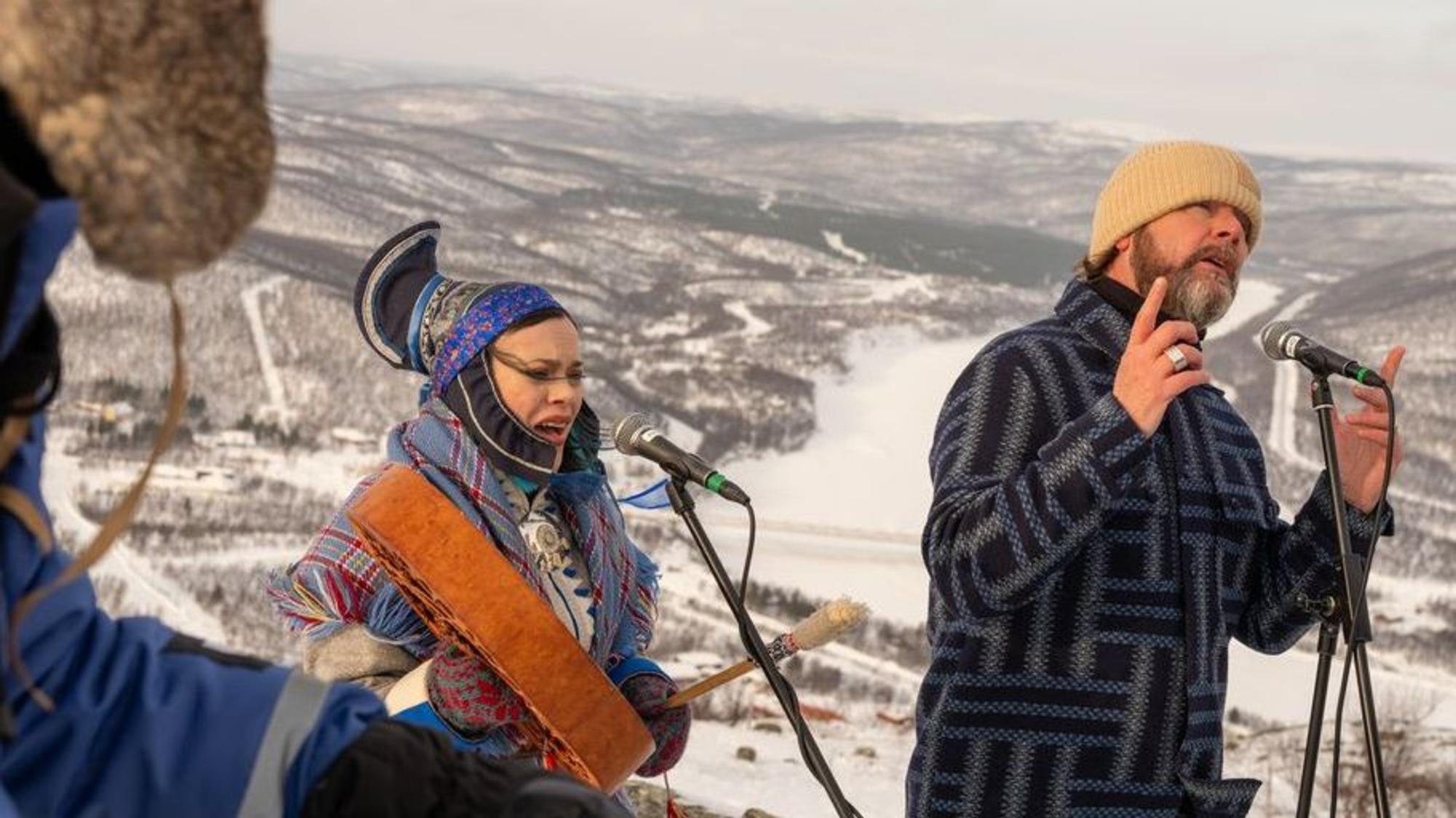 Eine Frau in nordischer Tracht und ein Man mit Mütze singen vor einer verschneiten Bergkulisse