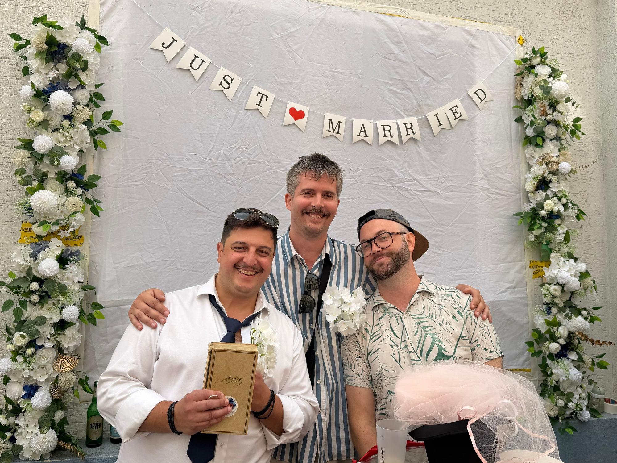 Drei Männer stehen unter einem mit Blumen geschmückten Schriftzug "Just Married".