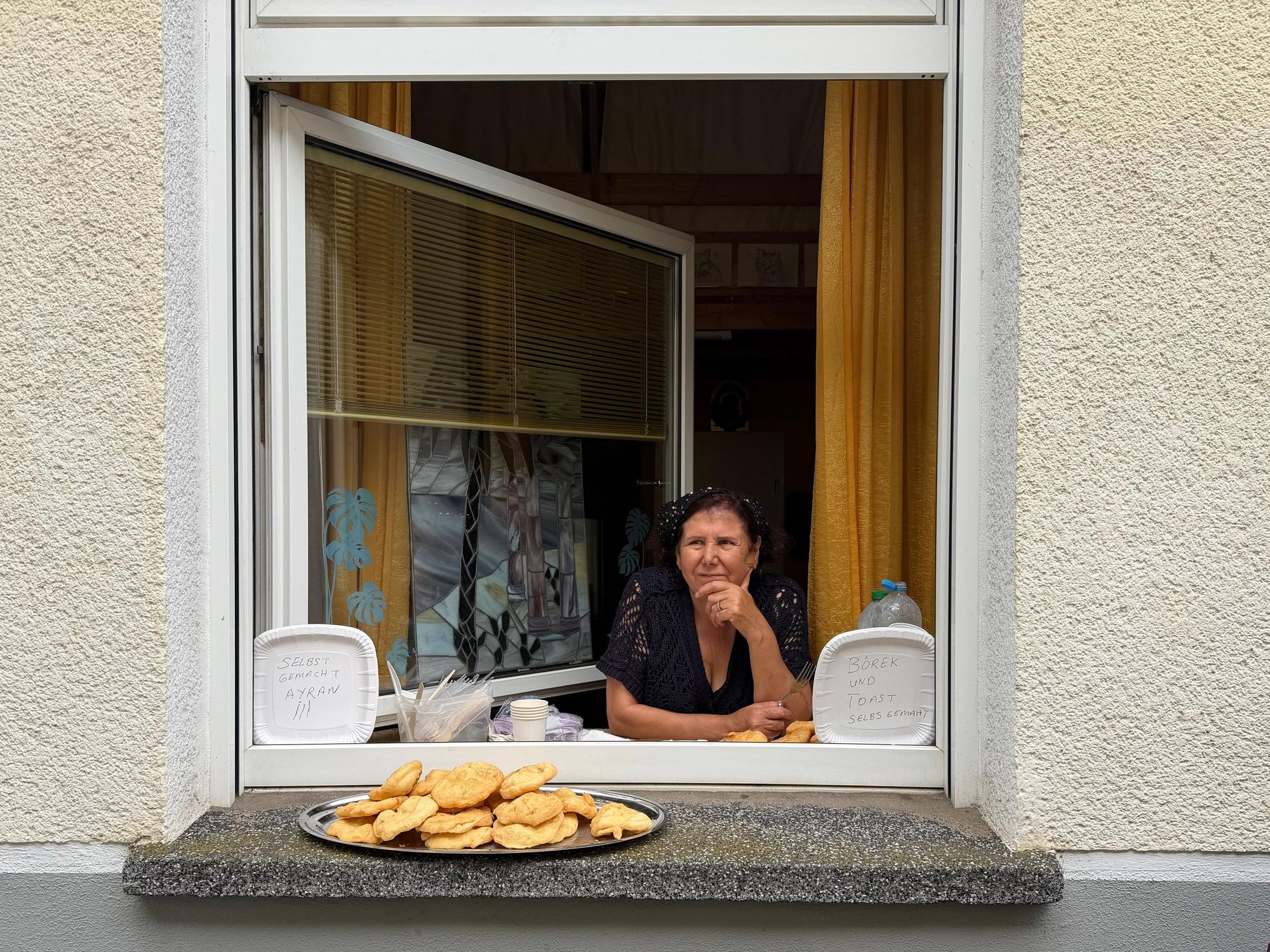 Eine Frau guckt aus ihrem geöffneten Wohnungsfenster, auf dem Fensterbrett steht ein Tablett mit Backwaren.