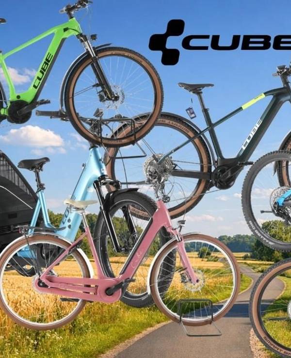 Cube E-Bikes, City Bikes, Mountainbikes, Lastenrad vor einer Landstraße im Grünen mit Bäumen, Wald und Wiesen im Hintergrund und blauem Himmel.