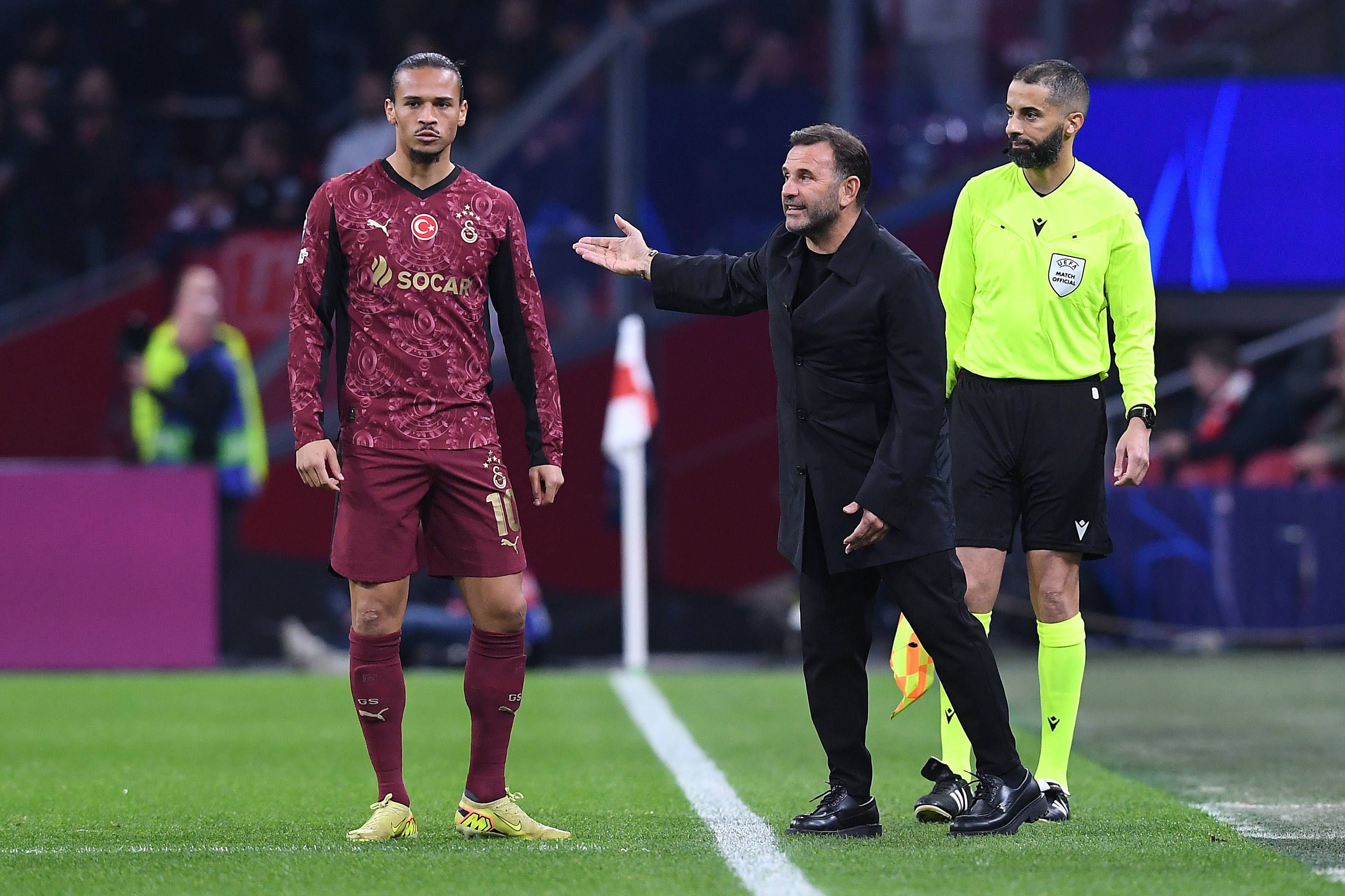 Leroy Sané steht an der Seitenlinie, neben ihm gestikuliert Trainer Okan Buruk.