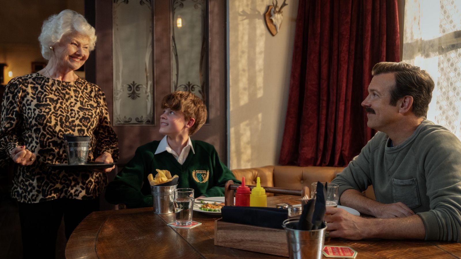 Die temperamentvolle Pub-Besitzerin Mae (Annette Badland) kümmert sich um Ted Lasso (Jason Sudeikis) und seinem Sohn Henry (Grant Feely). (Bild: © Apple TV / Collin Hutton)