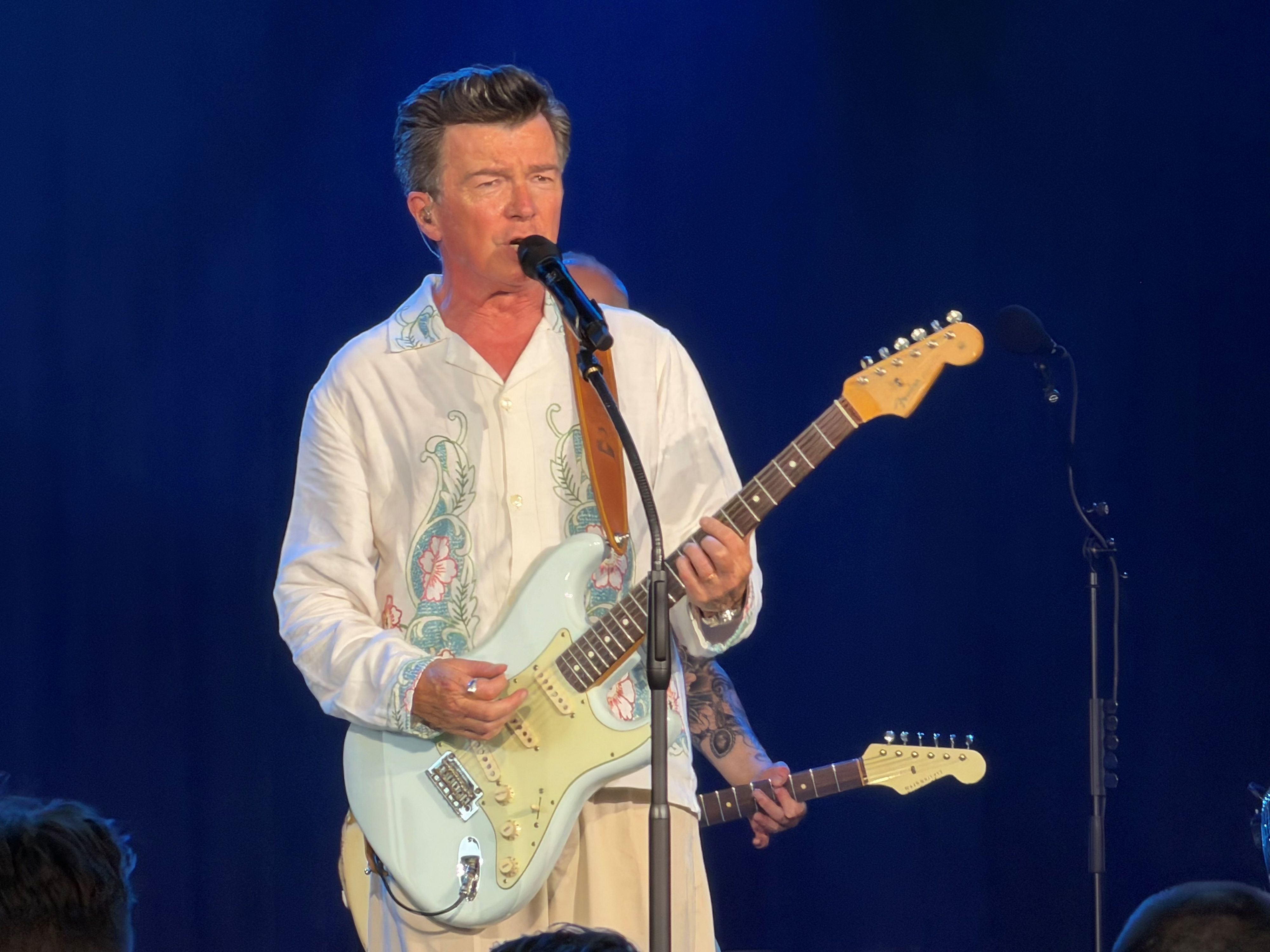 Rick Astley steht mit Gitarre auf der Bühne.