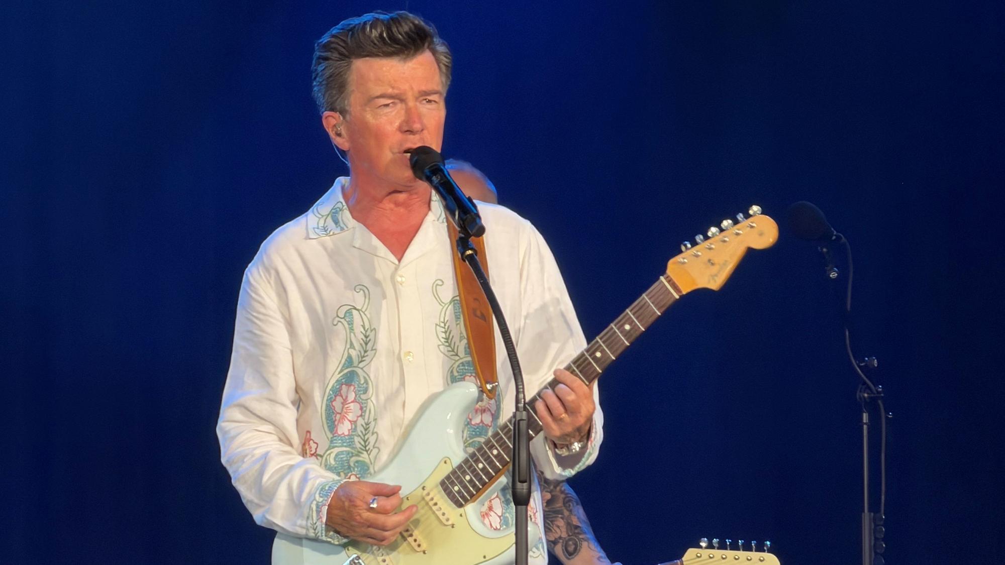 Rick Astley steht mit Gitarre auf der Bühne.