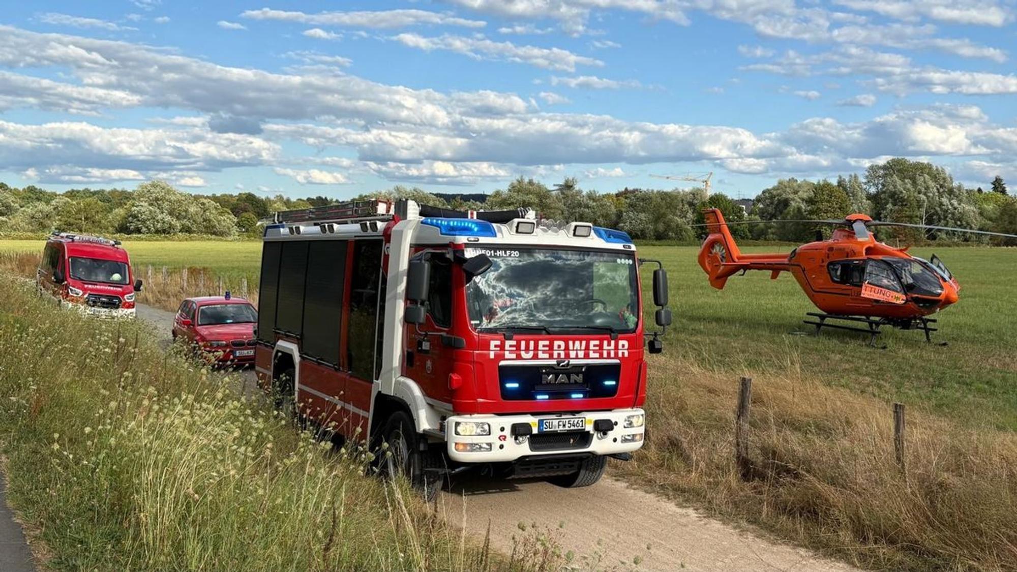 Auf einem unbefestigten Weg stehen Feuerwehrfahrzeuge, nebenan auf einem Feld ist ein Hubschrauber gelandet.