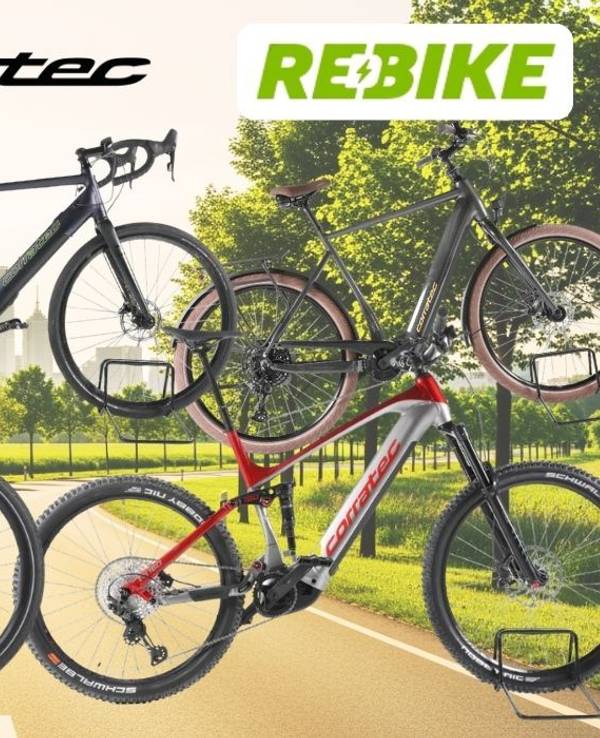 Mehrere Corratec E‑Bikes unterschiedlicher Typen – darunter Trekking-, City- und Mountainbikes – vor einer Landstraße an einem sonnigen Tag in Natur.