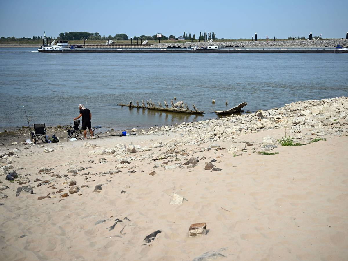 Ein Frachtschiff fährt bei Niedrigwasser auf dem Rhein an dem Wrack eines Bootes vorbei, das normalerweise unter Wasser liegt.